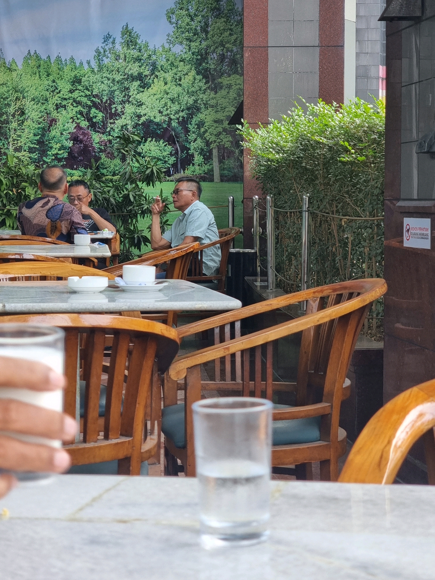 Three men are seated at tables outside a cafe in Central Jakarta, Indonesia, with a large mural of a forest as the backdrop. The man on the right is wearing a light blue collared shirt and glasses, and he is holding a cigarette to his mouth. The man in the middle is wearing a black t-shirt and glasses, and he is looking down at his table. The man on the left is wearing a patterned shirt and is facing away from the camera, talking to the man in the middle. Wooden chairs and marble-topped tables are arranged around the men. A glass of water sits on one of the tables in the foreground. In the background, a green hedge separates the seating area from a building with red and grey tiled walls. A sign with a prohibition symbol and text in Indonesian is visible on the building.