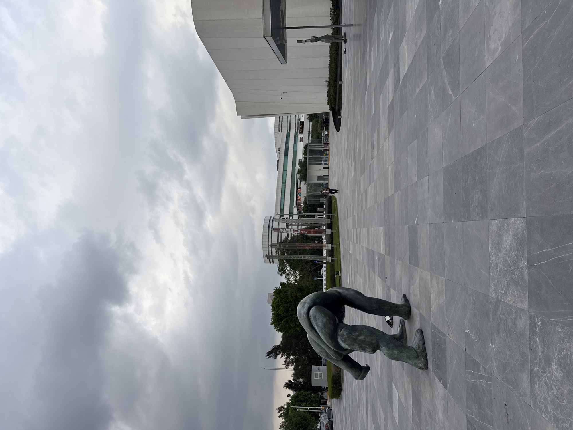 A bronze sculpture of two figures bent over is centrally located on a grey, tiled plaza in San Bernardino Tlaxcalancingo, Mexico. Behind the sculpture is a line of green trees, followed by a distant building with a cylindrical, latticed upper section. To the left, the sky is overcast with heavy, grey clouds. To the right is a large white building and another indistinct figure on the paved area in the distance.
