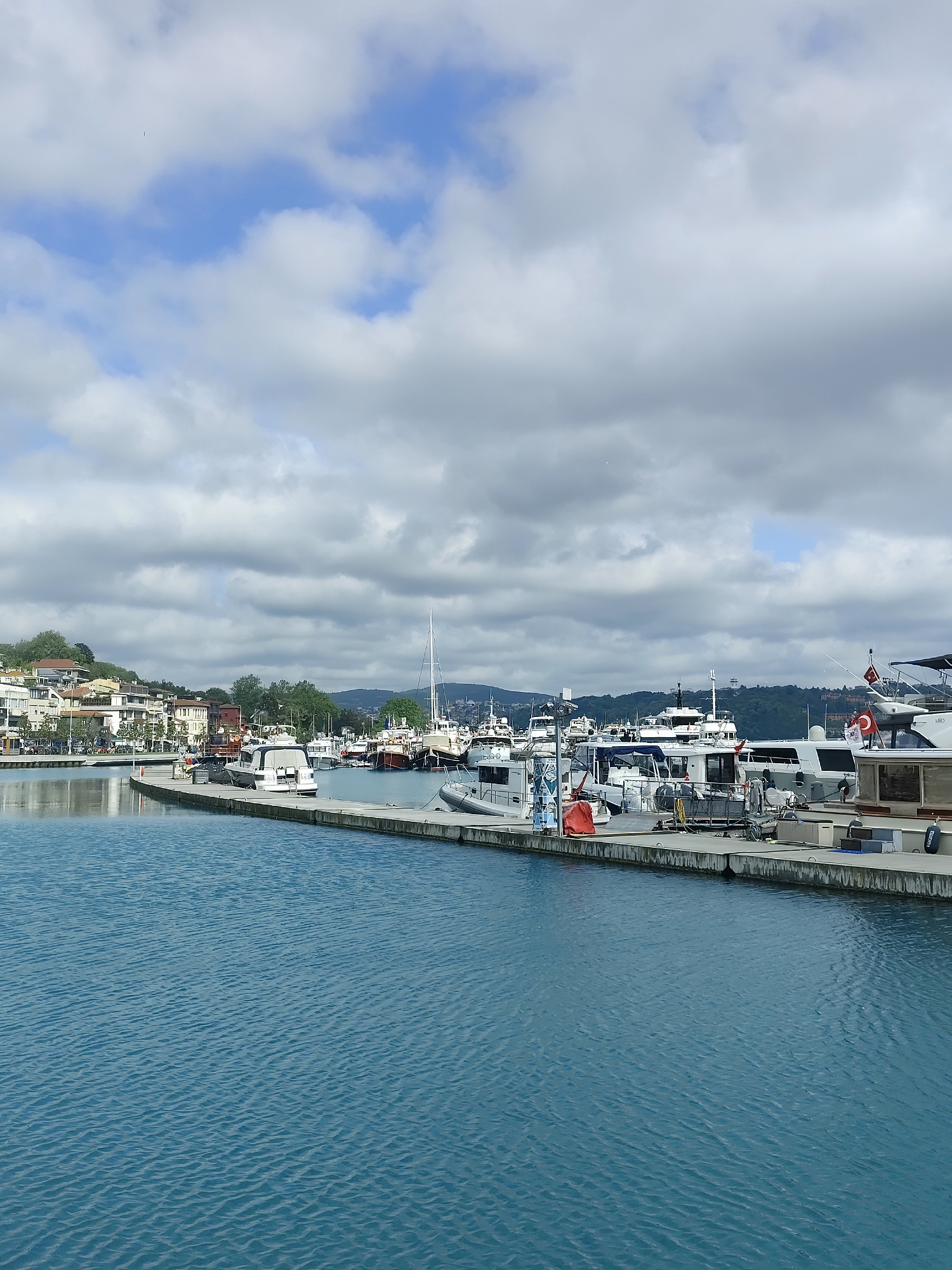 This image shows a marina in Sarıyer, Turkey. The water is a deep blue-green color, and there are many boats of various sizes docked along a concrete pier. The sky is cloudy with patches of blue. Buildings and trees are visible on the land in the background, along with signs of human activity. There are no people immediately visible in the photograph.