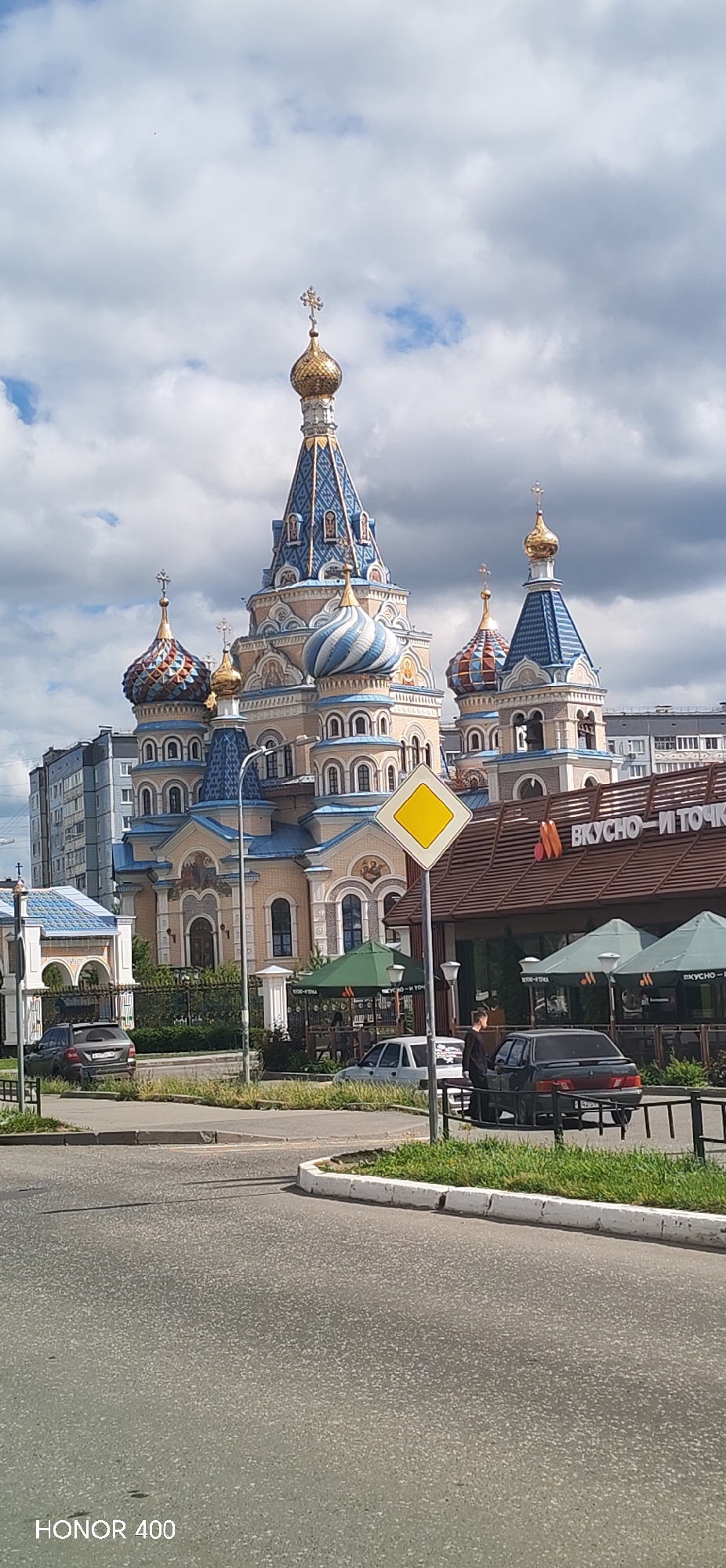 The image captures a view of the Transfiguration Cathedral in Izhevsk, Russia, on a cloudy day. The ornate cathedral, a prominent religious landmark, features multiple gold and blue-domed towers, characteristic of Russian Orthodox architecture. In the foreground, a street is visible with a road, sidewalk, and grassy areas. Several cars are parked or driving on the street. Adjacent to the cathedral is a building with awnings and signage that appears to be a food establishment, possibly "Vkusno — i tochka," a Russian fast-food chain. A yellow diamond-shaped road sign is prominently displayed in front of the establishment. The sky is filled with a mix of white and grey clouds, suggesting overcast weather. The overall scene depicts an urban environment with a blend of religious and commercial structures.