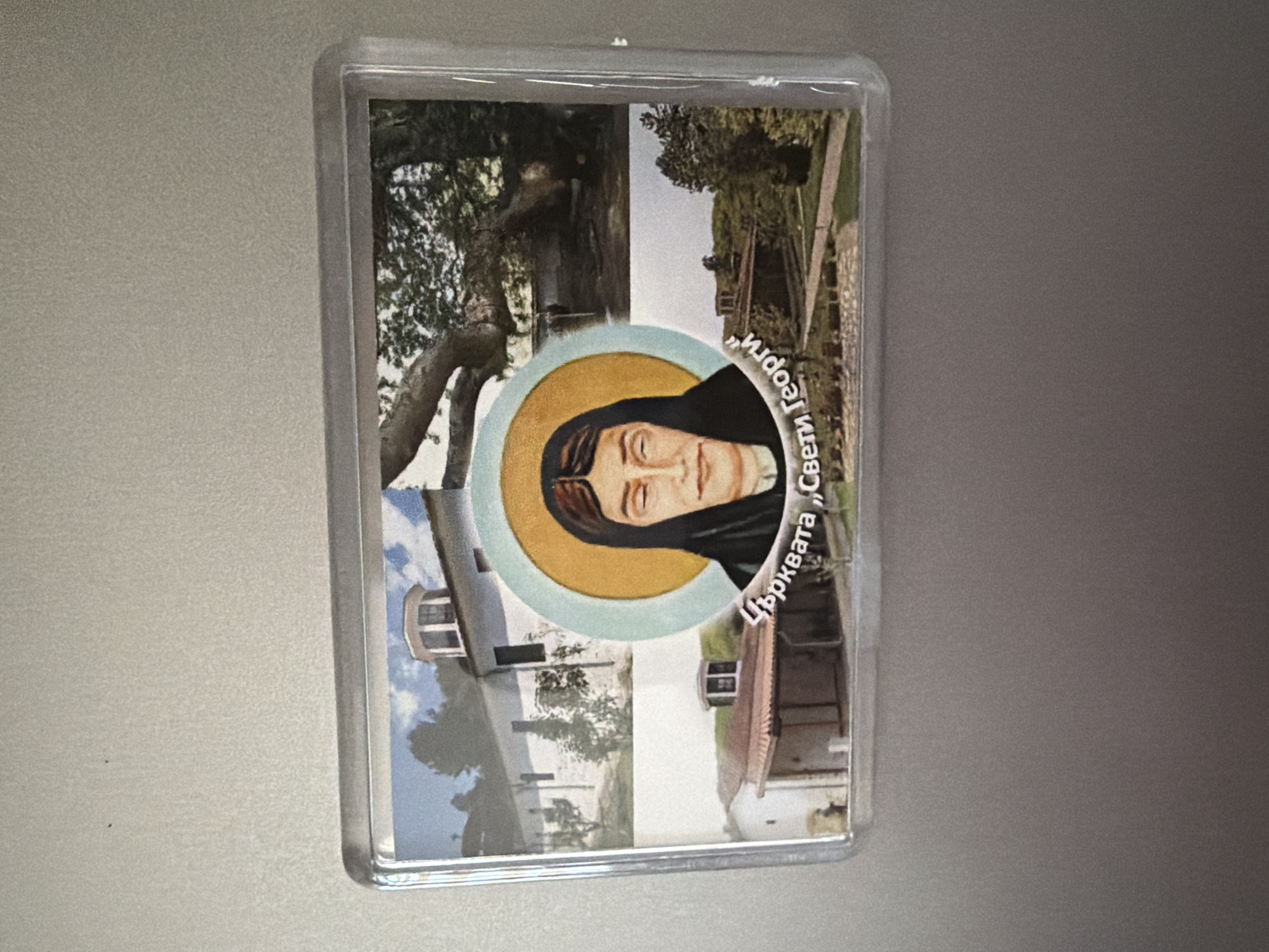 The image is a close-up of a rectangular magnet or similar souvenir item. It features a composite image with a portrait of a woman with dark hair, wearing a black veil and a yellow halo. She is looking downwards. The portrait is superimposed over a collage of three photographs. The top left photograph shows a white building with a dome, possibly a church or monastery, with trees and blue sky. The top right photograph shows a large, gnarled tree trunk and branches with dense foliage. The bottom photograph shows a smaller building with a reddish-brown tiled roof, and a paved walkway or courtyard in front. The text "Църквата „Свети Георги“" is visible in Cyrillic script, which translates to "Church 'Saint George'". This likely indicates that the souvenir depicts the Church of Saint George in Pazardzhik, Bulgaria. The magnet is clear and appears to be in good condition, with no visible damage.
