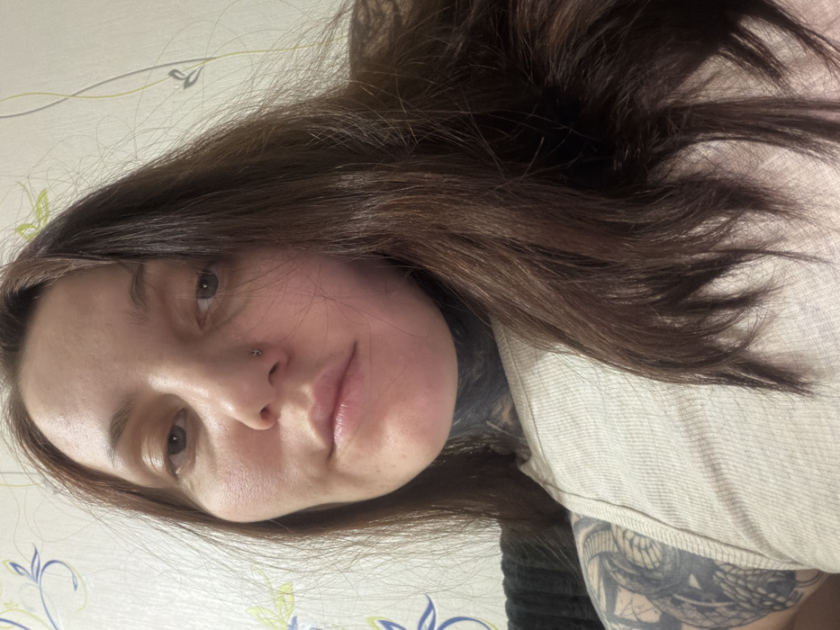 A woman with long brown hair gazes into the camera. Her skin appears smooth and fair. She has a nose piercing and full lips. Visible tattoos mark her neck and arm. She wears a light-colored top. The background is a wall with a subtle floral pattern. The perspective is an unusual angle, possibly a selfie taken from above.