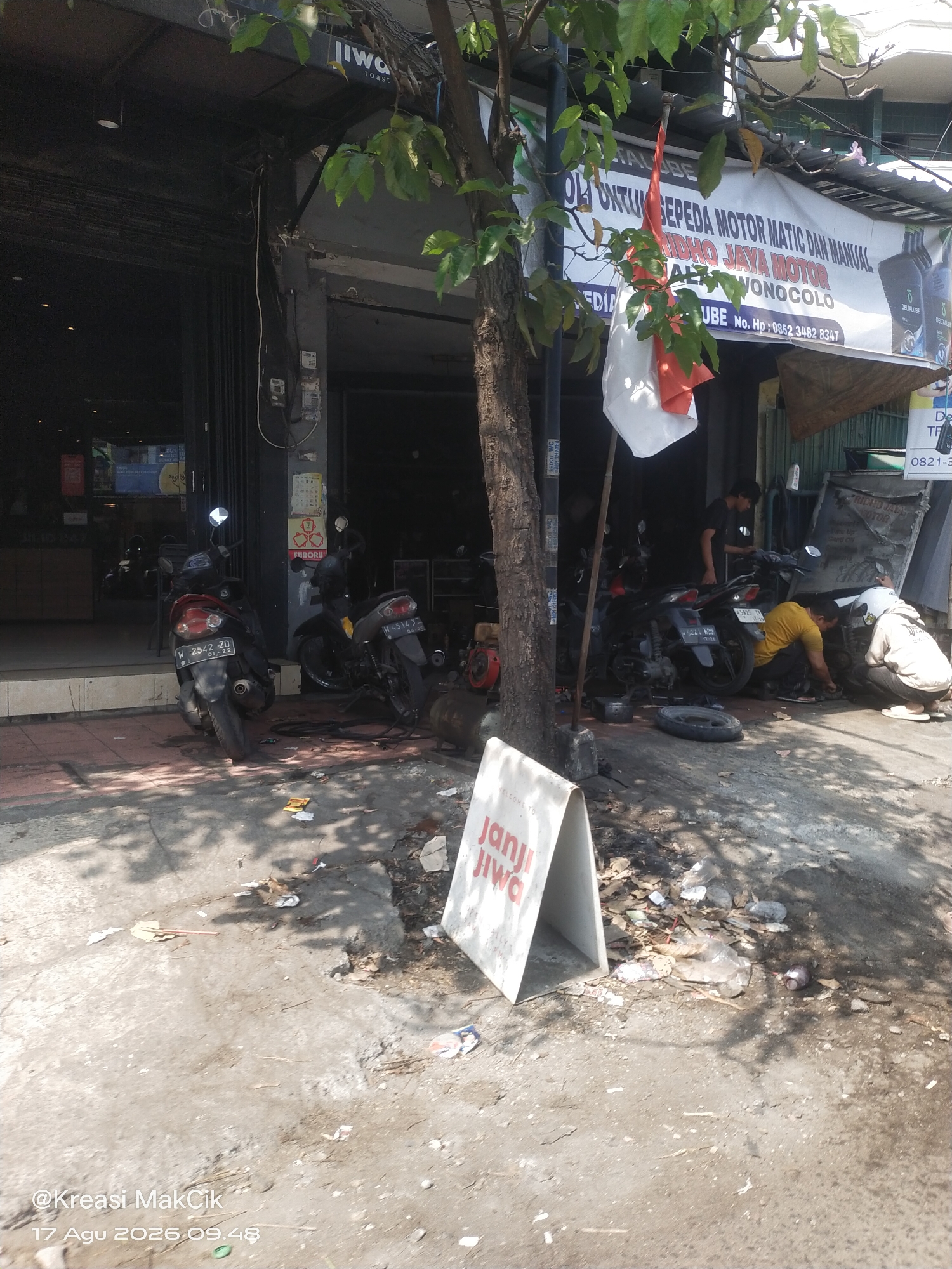 In the city of Wonocolo, Indonesia, a motorbike repair shop is captured on a sunny day. The shop front, with its open entrance, reveals a workspace cluttered with several motorcycles in various states of repair. Two mechanics are actively working on a motorcycle, one in a yellow t-shirt and the other in a light-colored hoodie and helmet. Another individual, wearing a black t-shirt, stands in the background near a motorcycle.
In front of the shop, two motorcycles are parked on the sidewalk. A white A-frame sign with red text that reads "JANJI JIWA" is placed near the entrance, partially obscured by a tree and foliage. The sidewalk area in front of the shop is dirty and littered with debris, including plastic bags, wrappers, and small stones. The shop itself has a dark awning overhead with some signage visible. The overall environment suggests a busy, functional repair shop operating in an urban setting. The image also includes a timestamp and username in the bottom left corner: "@Kreasi MakCik 17 Agu 2026 09.48".