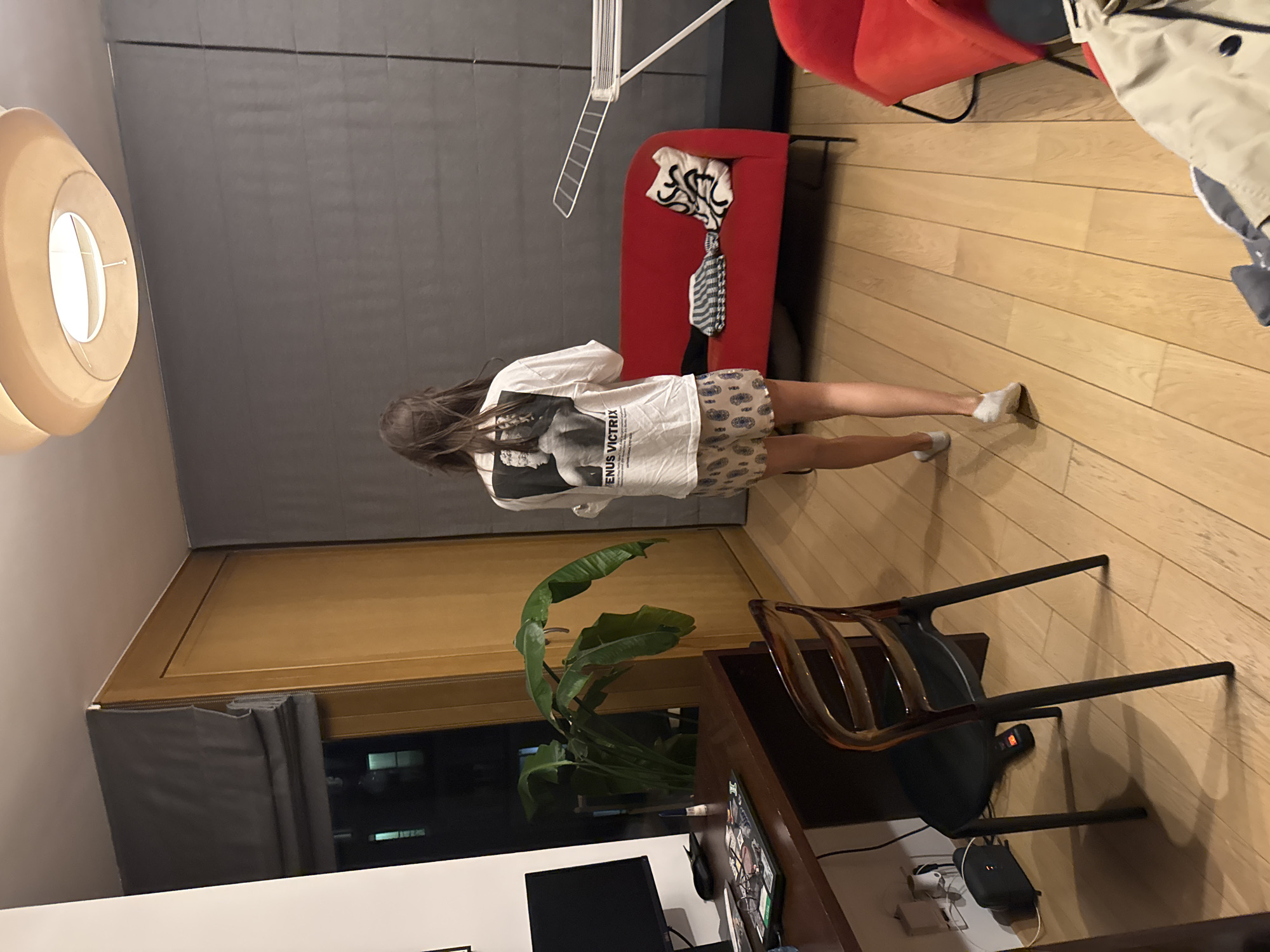 A medium shot captures the interior of an apartment. A young woman with long, dark hair stands near a red sofa, facing away from the camera. She wears a white T-shirt with a black-and-white graphic and patterned shorts. A clothing rack with drying clothes stands between her and the sofa.
The apartment features light wood floors and a dark gray wall. A large, round, beige lamp fixture hangs from the ceiling on the left. A wooden desk with a computer monitor, a small plant, and a wooden chair is visible in the foreground. A window with dark gray curtains is located behind the desk.
The scene is likely set in Warsaw, Poland, but there are no visible landmarks to confirm this. The image depicts a casual, everyday scene within a modern apartment interior.