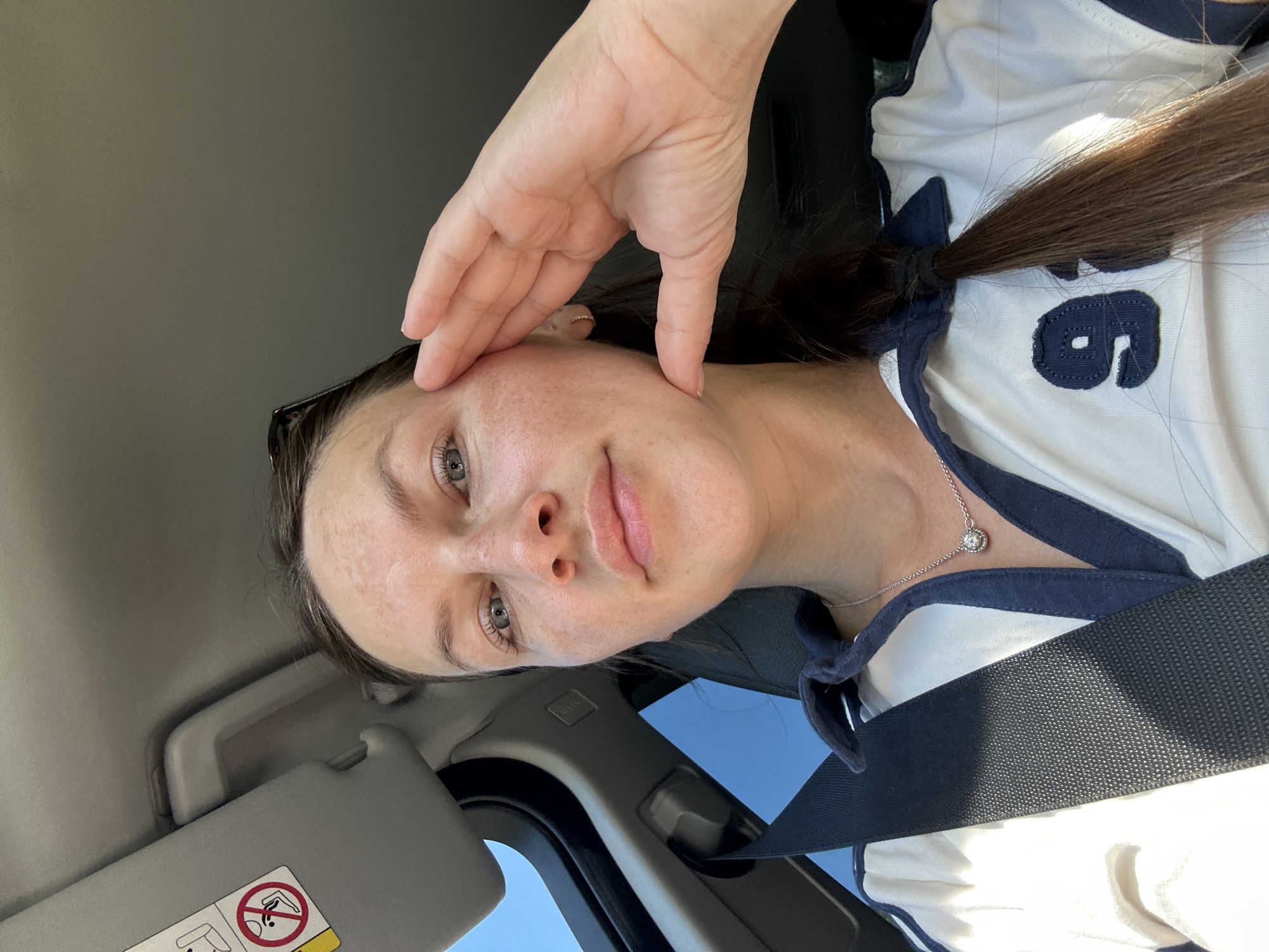 A woman is taking a selfie in a car. She is wearing a white polo shirt with blue trim and a number 9 on the front. She has on a silver necklace with a pendant. Her hand is resting on her face. The sun visor in the car is down, and there is a seatbelt visible. The background shows a clear blue sky and part of the car's interior. The image was taken in Vrela, Montenegro.