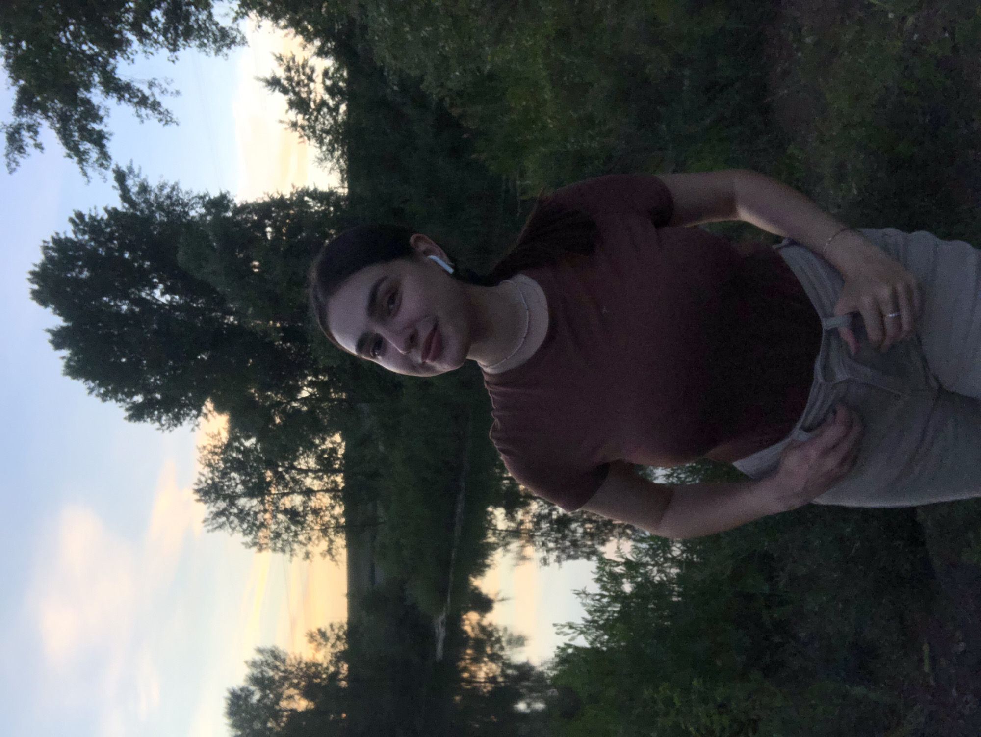 A young woman with dark hair pulled into a ponytail, wearing a brown t-shirt and light-colored pants, stands outdoors near a body of water during sunset. She has on white wireless earbuds and a delicate necklace. Her left hand is placed on her pants pocket, and a ring is visible on her left ring finger. The background features a calm body of water reflecting the sky, lush green foliage, and trees silhouetted against a sky with hues of orange, pink, and blue, indicative of a sunset. The location is the embankment of the Kama River in Chaikovsky, Russia.