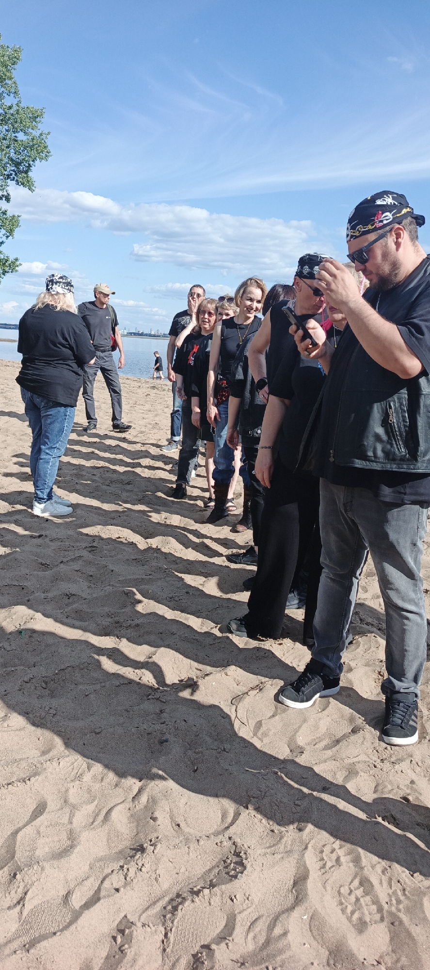 A group of approximately 10-12 adults is gathered on a sandy beach next to a large body of water under a clear blue sky with scattered white clouds. The setting is an outdoor, daylight environment, with long shadows cast across the sand, suggesting late morning or early afternoon. In the background, across the body of water, distant industrial or urban structures are visible on the horizon. A tree with green foliage is partially visible on the far left.
The individuals are primarily dressed in dark clothing, including black t-shirts, jeans, and vests. Several people wear patterned bandanas (some with skull motifs) and sunglasses. On the far left, a person with light-colored hair, wearing a black t-shirt, blue jeans, and white sneakers, stands facing right, appearing to converse with another male figure in a beige cap, dark t-shirt, and dark trousers, who has a red bag slung over his shoulder.
To the right, a line of people extends towards the left. The individual closest to the camera on the right is a bearded male wearing sunglasses, a black bandana with white and red patterns, a black t-shirt, and a black leather vest; he is actively engaged with a smartphone held in both hands. Adjacent to him in the line are other individuals, including a person also wearing a black bandana and sunglasses, a blonde female smiling towards the camera, and a female with glasses wearing a black t-shirt with a red star design. A small child or person is visible in the shallow water further along the beach in the distant background. The sand shows various footprints and undulations. The image provides no specific visual cues to identify the location as Noviy, Russia.