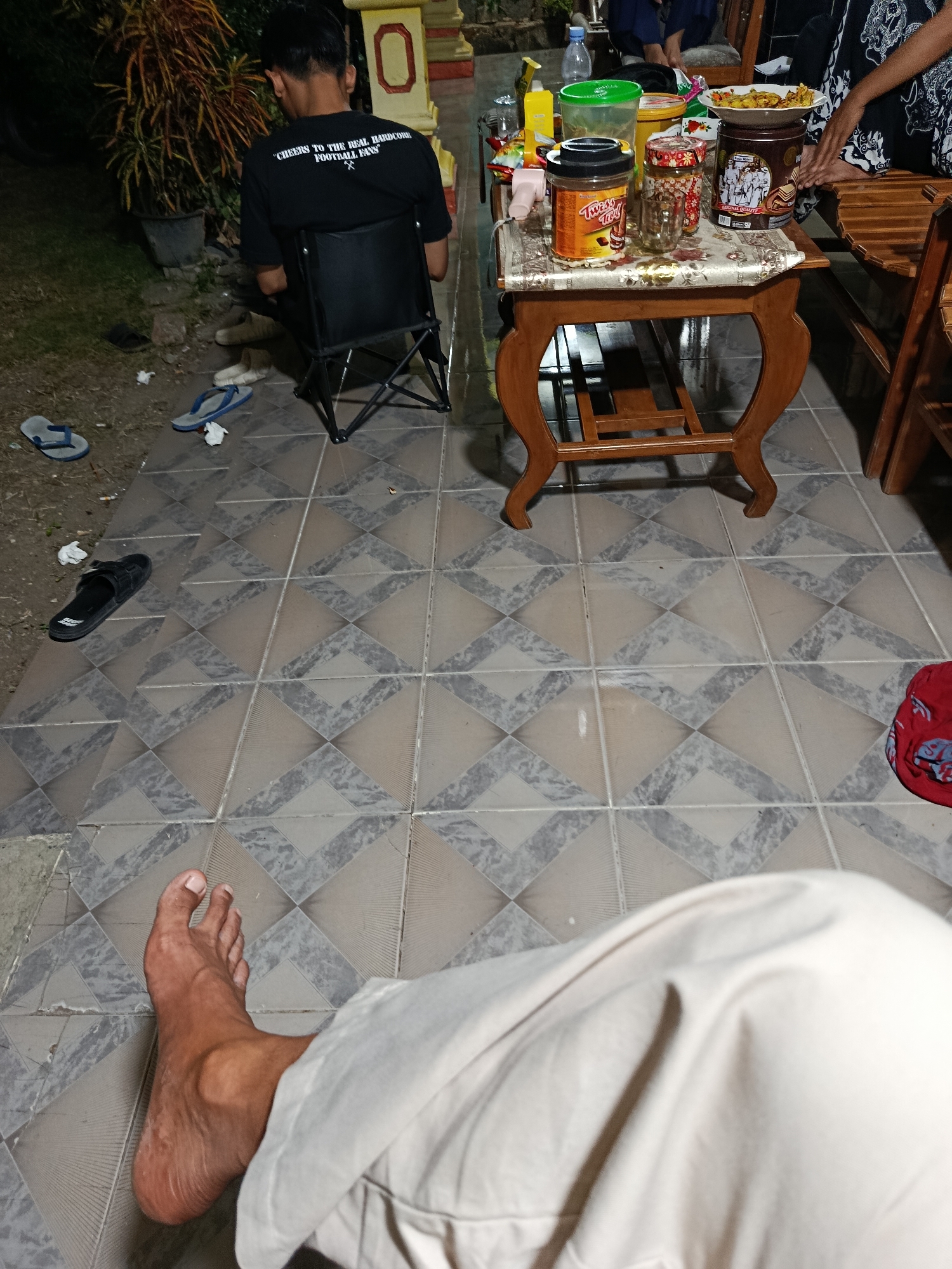The image depicts an outdoor scene at night, likely in Sarwadadi, Indonesia. In the foreground, a person's bare foot, clad in light-colored pants, rests on tiled flooring. The tiles are arranged in a diamond pattern with alternating beige and gray tones. To the left, a black flip-flop rests on dirt. Further left, two pairs of blue flip-flops are scattered on the dirt ground, with some white paper debris near them.
In the mid-ground, a dark-colored folding chair is occupied by a person wearing a black t-shirt with white text that reads "CHEERS TO THE REAL HARDCORE FOOTBALL FANS". Behind them, a plant with long, colorful leaves in a black pot is visible.
The background features a low wooden table with intricately carved legs, covered by a patterned tablecloth. On the table, various items are present, including a plate of food, several jars and containers of food products, a bottle of water, and a yellow spray bottle. To the right of the table, a wooden bench and chairs are partially visible. Another person is seated on the bench. The overall lighting suggests an outdoor gathering after dark.
