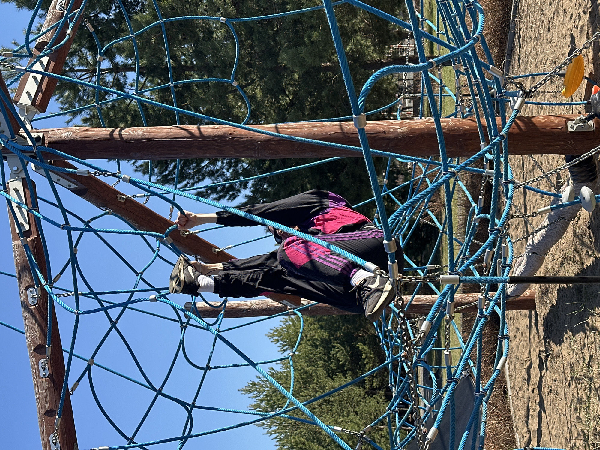 A person is climbing a blue rope net structure at a playground in Русский Вожой, Russia. The person is wearing black pants and a black jacket with pink stripes. The playground has a wooden support structure and a tan dirt surface. Trees are visible in the background under a bright blue sky.