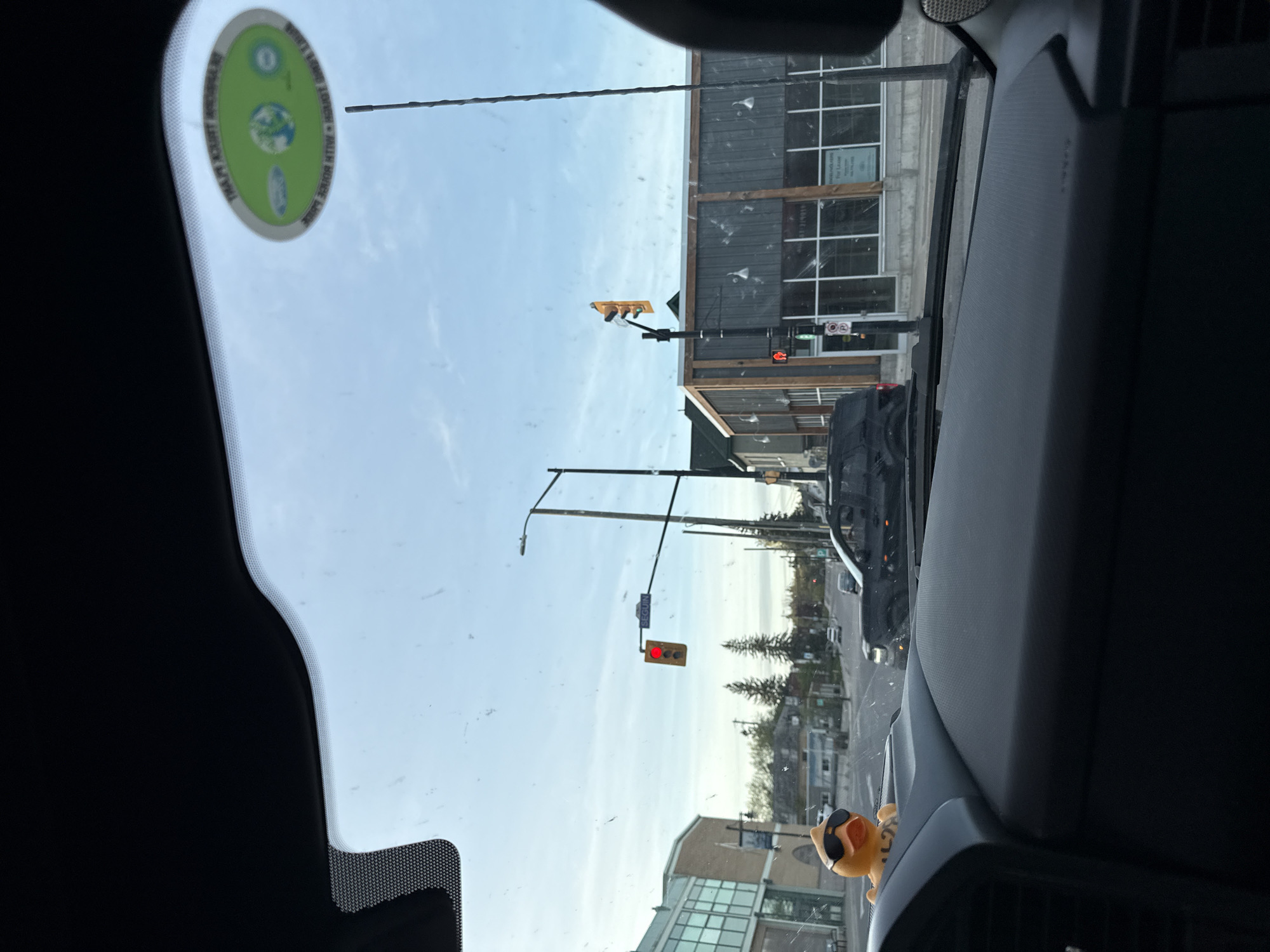 The image is taken from inside a vehicle, looking out through the windshield at a street scene. A green sticker is affixed to the windshield. Outside, there is a traffic light showing red. A sign reads "SEVERN." Across the street, there's a building with a gray facade and large windows. In the foreground, on the car's dashboard, is a small toy character wearing sunglasses. The sky is bright and clear.