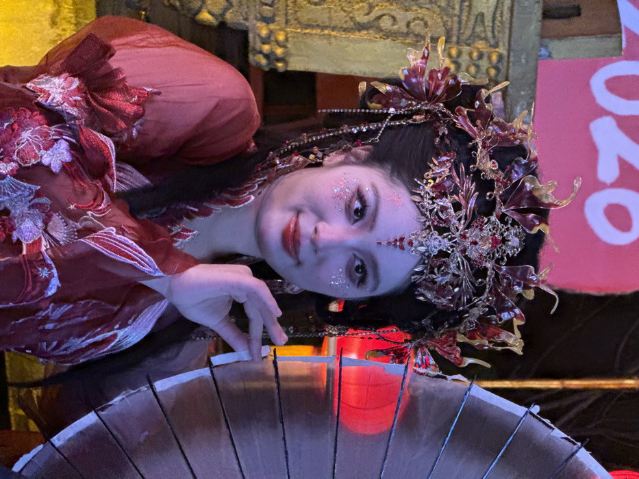 An individual, identified as a female, is depicted in a medium-shot portrait, captured within Yuzhong District, China. She is positioned slightly to the left of the frame's center, making direct eye contact with the viewer. The subject wears an elaborate, reddish-maroon traditional costume, featuring intricate floral embroidery in lighter red and silver/white threads on the visible sleeve and bodice. A large, ornate headpiece adorns her dark hair, styled in an updo; the headpiece is gold-toned with red gemstone accents and dangling beadwork. Her face is made up with glitter around the eyes, dark eyeliner, and glossy red lipstick; a red gem-like application is present on her forehead. Her right hand (viewer's left) gently grips the edge of a partially visible, dark-colored traditional parasol or umbrella, which occupies the right side of the frame. In the background, a large red sign with partial white characters, possibly "2026", is visible on the upper left. Blurred red spherical objects, consistent with Chinese lanterns, are discernable behind the parasol on the right. The scene is illuminated by artificial warm light, suggesting an evening or indoor setting, likely a cultural event or performance.