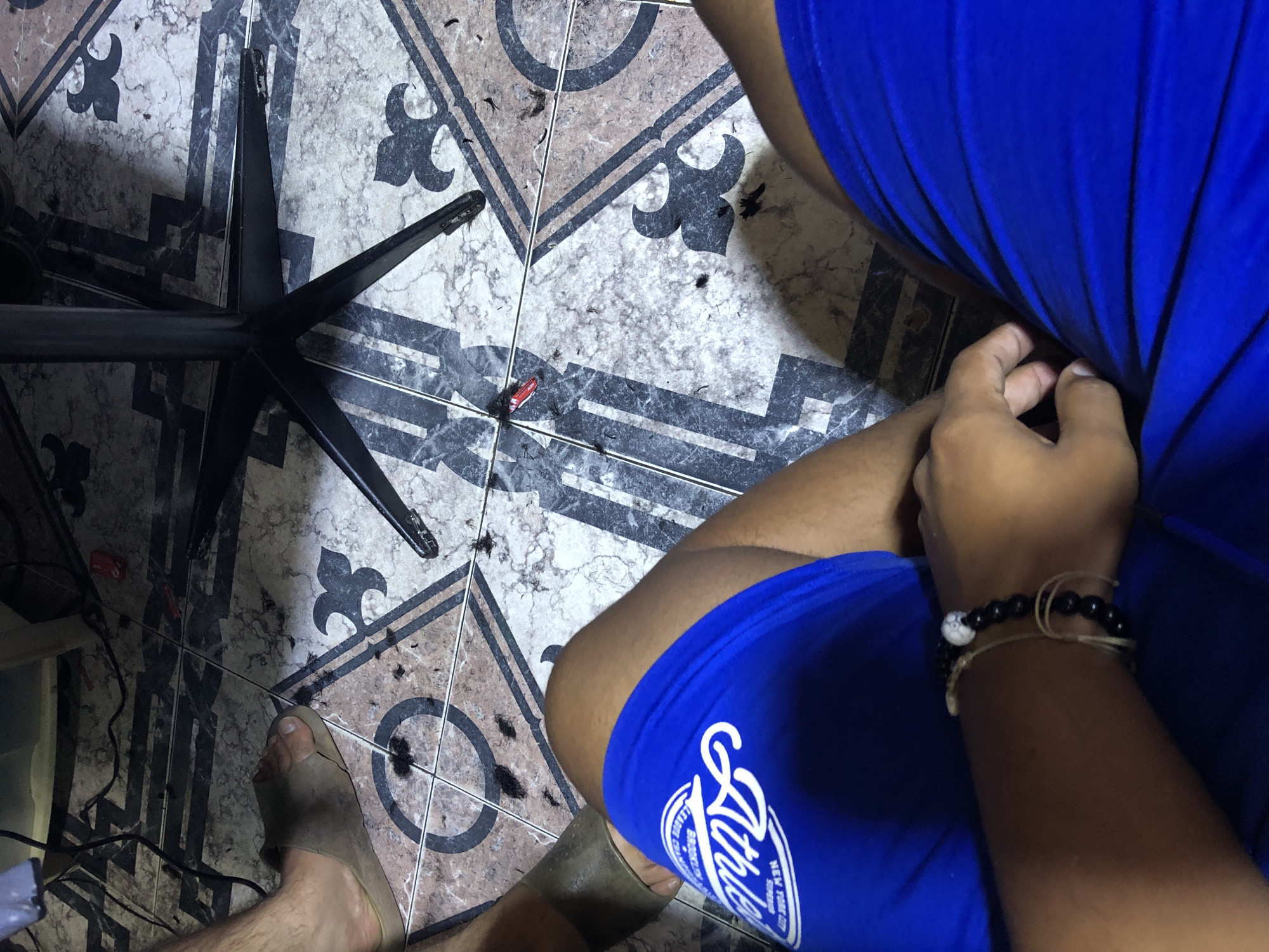 A high-angle shot depicts a person sitting on a chair, presumably in a home. The person is wearing blue athletic shorts and a bracelet. The floor is tiled and covered in hair clippings, suggesting a recent haircut. A black chair base is visible in the lower left. A red tube is on the floor near the chair base. The location is presumed to be Altagracia, Venezuela.