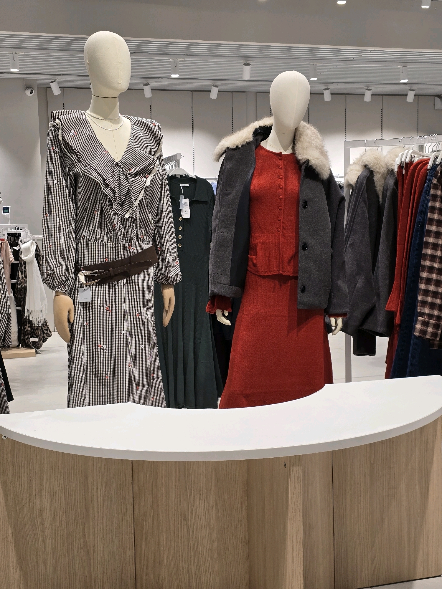 The image shows two mannequins displaying outfits in what appears to be a clothing store. The mannequin on the left is wearing a long-sleeved, collared dress with a black and white checkered pattern and red floral accents. The dress has ruffles around the neckline and down the front. It is cinched at the waist with a dark brown belt. The mannequin on the right is wearing a red knitted outfit consisting of a top with a high neckline and a long skirt. Over the red outfit, it wears a dark gray coat with a light brown fur collar. Several other garments are visible on racks in the background, including a dark green dress, a plaid jacket, and red and blue items of clothing. The store has bright overhead lighting and white ceiling panels. The foreground features a curved white countertop with a light wood-grained base. This scene is located in the city of Khimki, Russia.