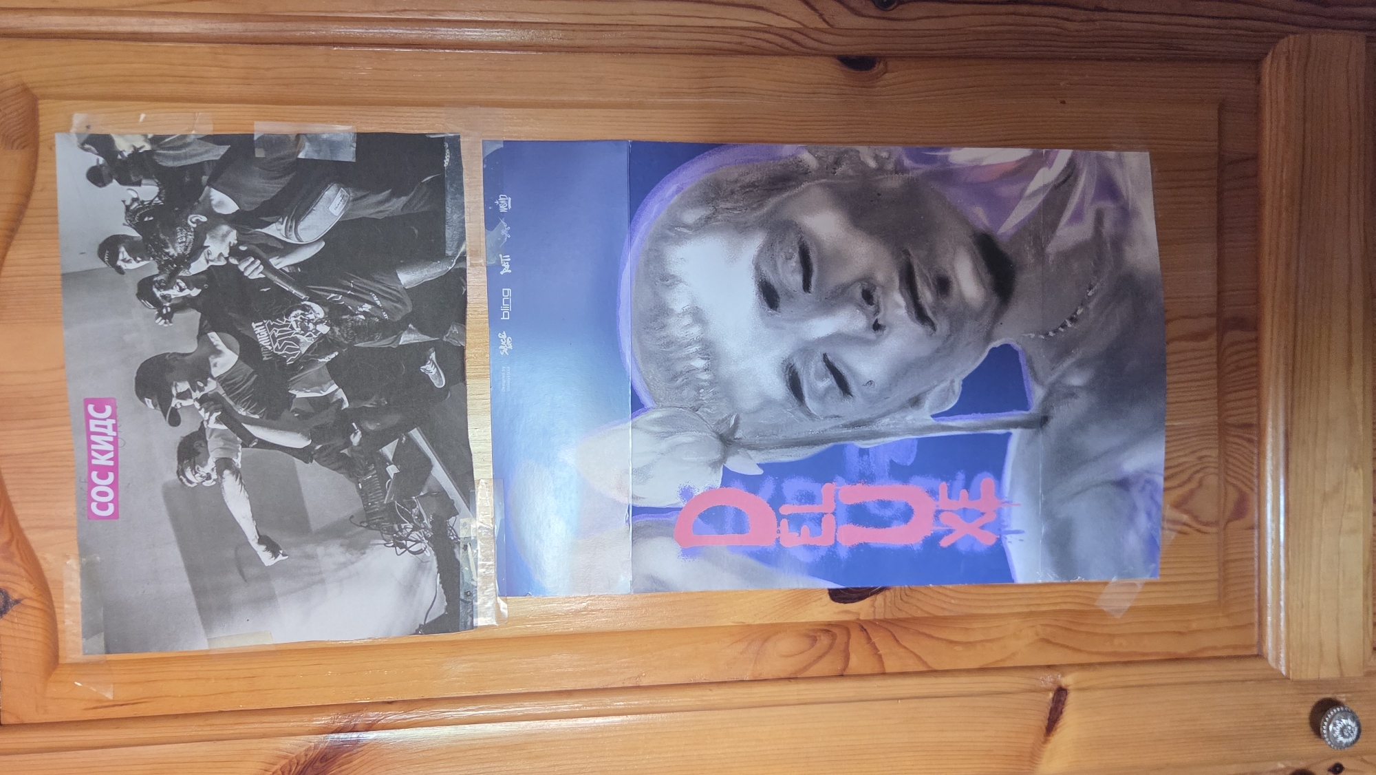 The image shows two posters attached to a wooden surface, likely a door or a cabinet, using clear tape. The poster on the left features a black and white image of a band performing on stage, with several members visible. The text "COC КИДС" is printed vertically on the left side of the poster. The poster on the right has a stylized image of a person's face in a silver or metallic tone against a blue background. Pink text is also visible on this poster. The wooden surface has a natural wood grain and is likely indoors. The location is not determinable based on the visual information provided.