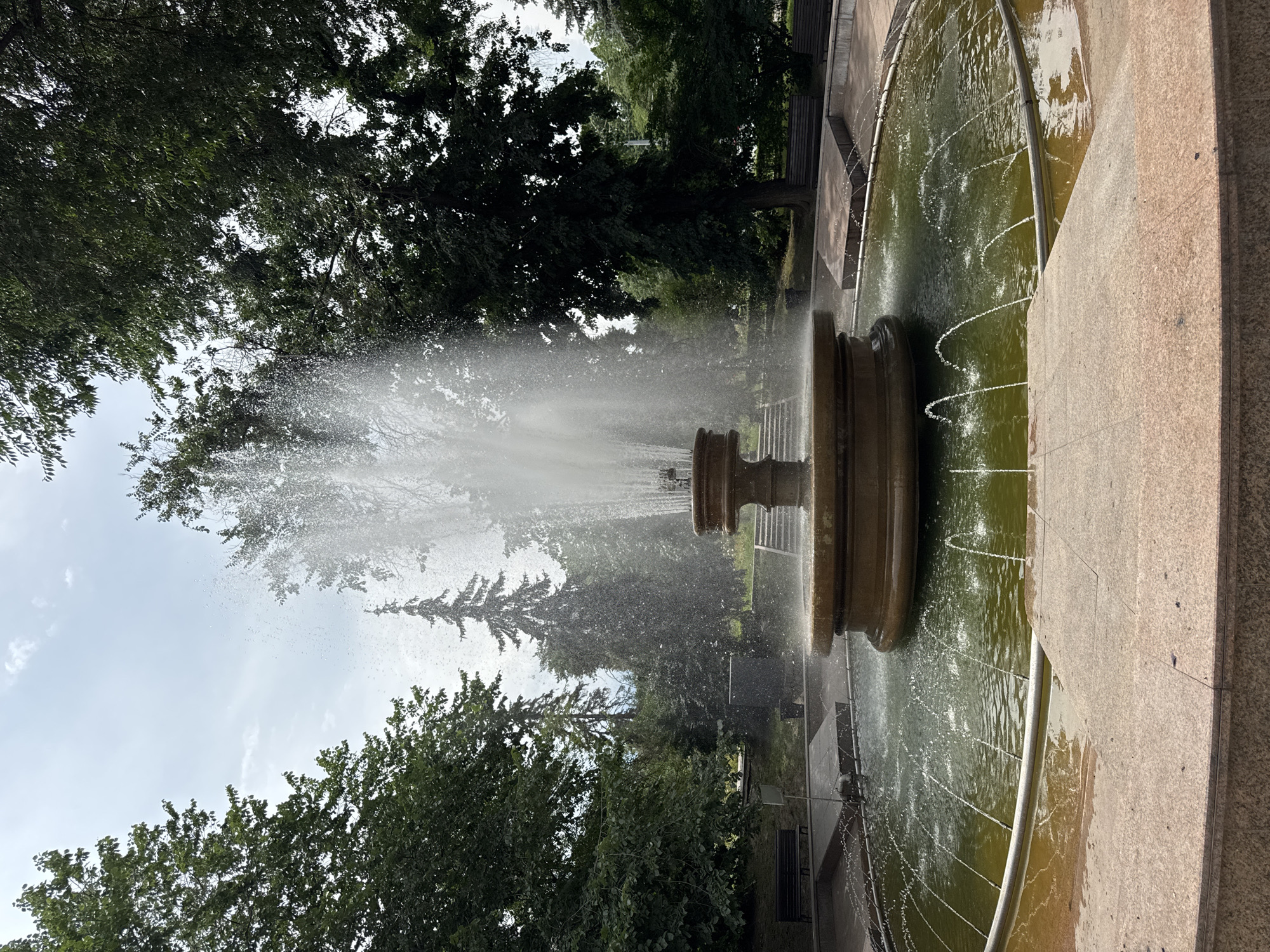 The image depicts a large, multi-tiered fountain in a park setting. The fountain has a broad, shallow basin filled with green water, from which numerous smaller jets of water arc outwards. In the center, a taller, more substantial section of the fountain sprays a powerful column of water upwards and outwards, creating a misty effect. The fountain is surrounded by a stone ledge and walkway, beyond which are pathways, park benches, and lush green trees. In the background, there are more trees, including a prominent evergreen, and a cloudy sky. The scene appears to be a public park in Almaty, Kazakhstan, likely a recreational area. There are no people visible in the frame.