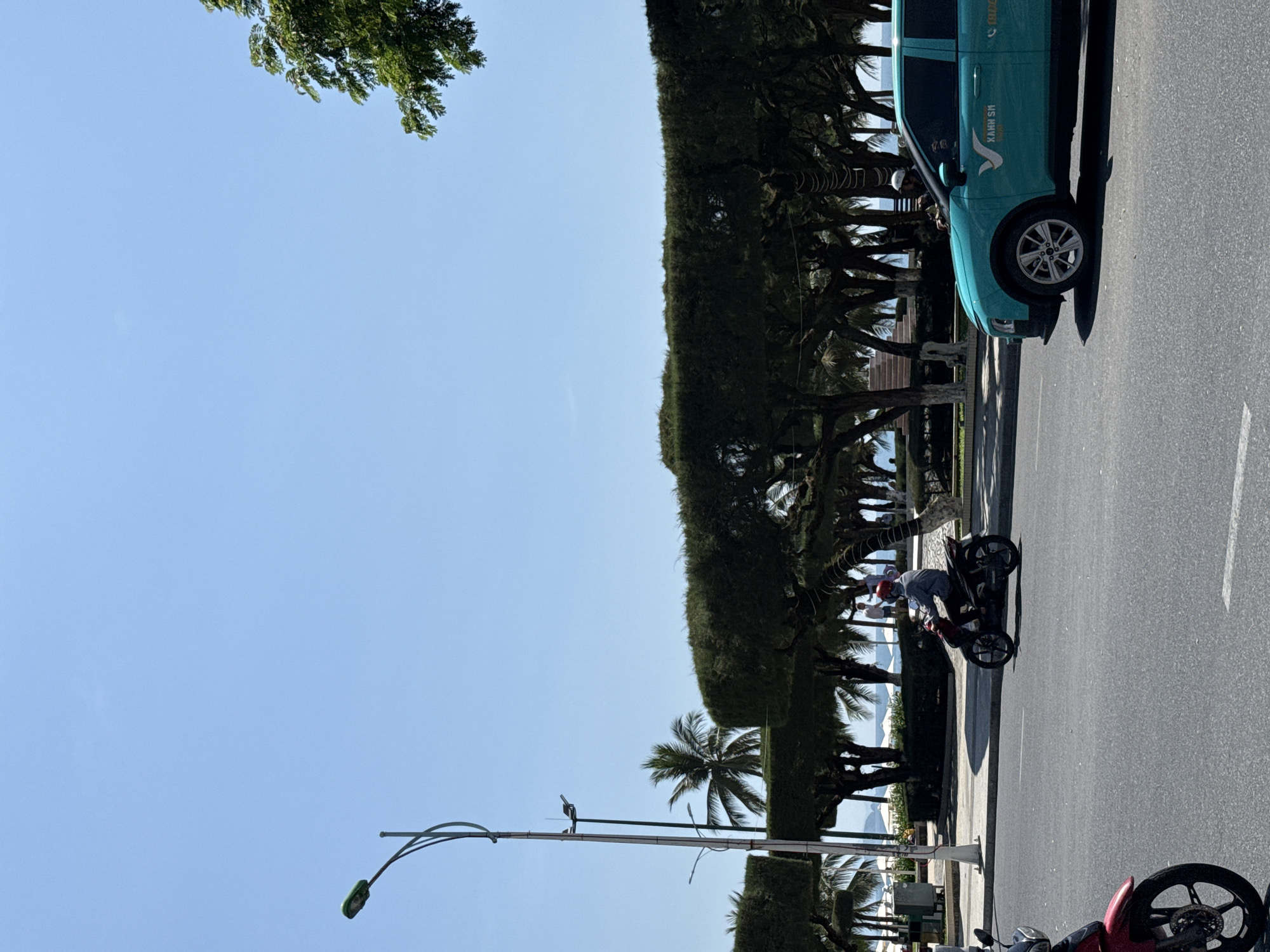 A street scene in Nha Trang Ward, Vietnam, shows a bright, sunny day with a clear blue sky. In the foreground, a gray asphalt road is visible with a white lane marker. A teal-colored car, identified as a XANH SM TAXI, is driving on the road. To the left, a red motorcycle is partially visible. Further down the road, a person wearing a blue shirt and red helmet is riding a dark-colored motorcycle. Beyond the road, a row of neatly trimmed, dark green trees forms a hedge-like barrier. Behind the trees, palm trees and the ocean are visible. A tall lamppost stands on the left side of the image.
