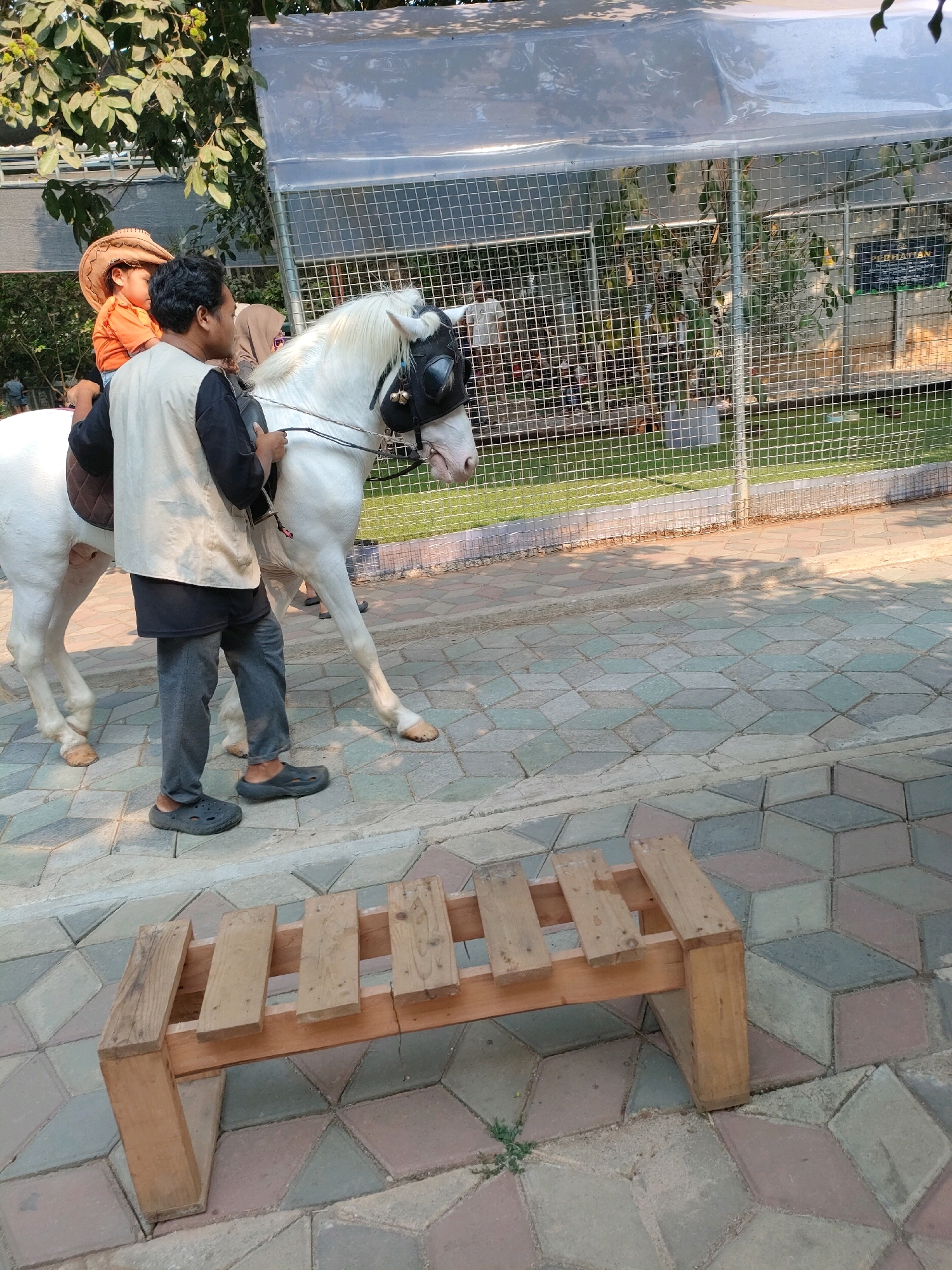 A white horse with a black bridle and blinders is being led on a patterned paver pathway by a man wearing a vest over a black shirt and dark pants. On the horse's back is a child wearing an orange shirt and a cowboy hat. Behind the horse, another person, possibly a woman, is partially visible wearing a headscarf. The horse and its riders are moving from left to right across the frame. To the right of the pathway is a fenced enclosure with green grass and trees visible inside. In the foreground, a small, rustic wooden bench made of planks is placed on the pathway. The lighting suggests it is daytime, possibly late afternoon, with dappled shadows falling on the ground from overhead foliage. The scene appears to be an outdoor recreational area, possibly a park or a petting zoo in Cileduk, Indonesia.