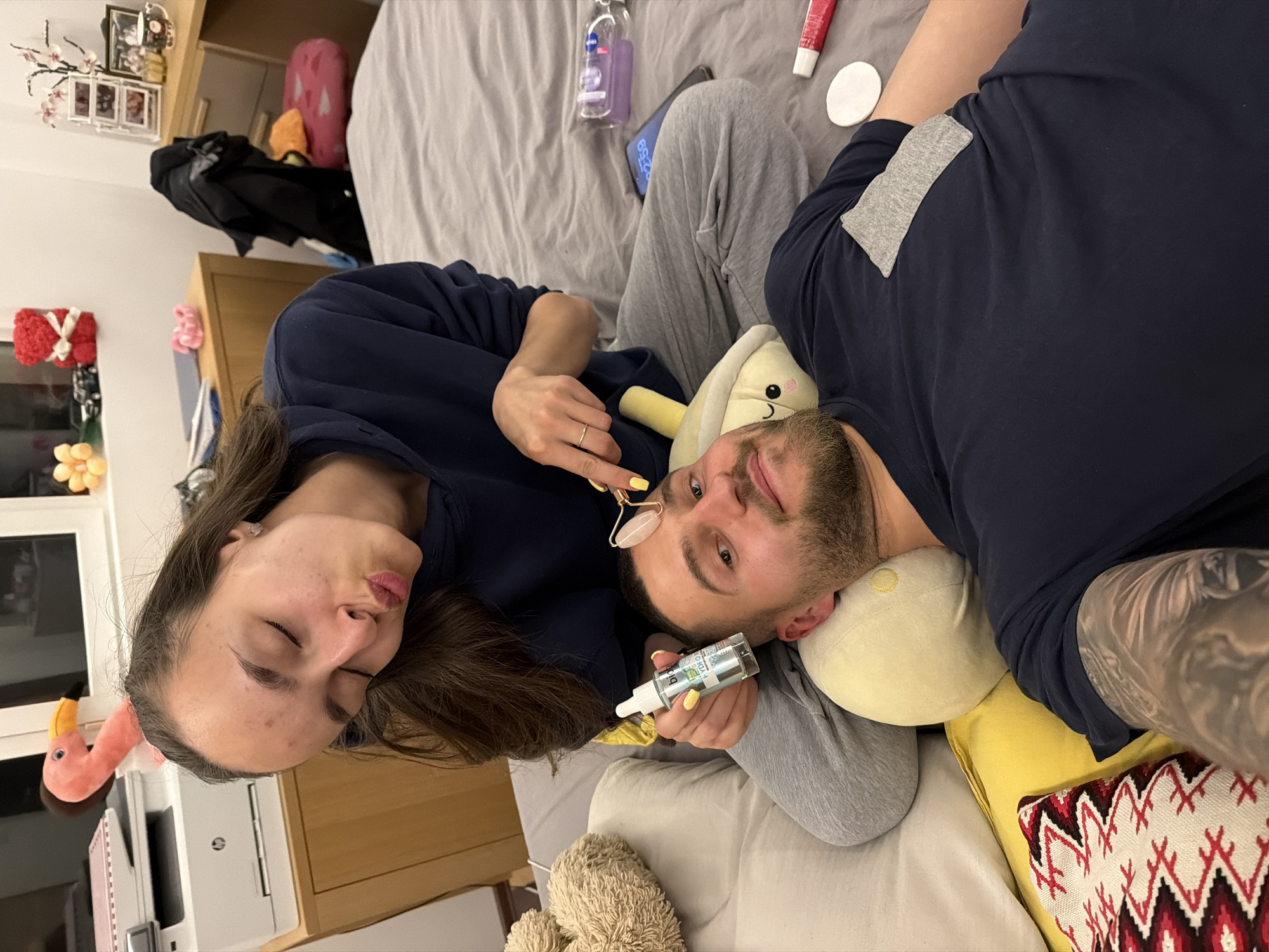 The image shows a couple lying on a bed indoors. The woman is positioned to the left, wearing a dark blue shirt, with long brown hair. She is holding a facial roller. The man is positioned to the right, wearing a navy shirt. He is holding a dropper bottle. They are lying on a gray sheet with a number of bottles. The location of this scene is not discernible.