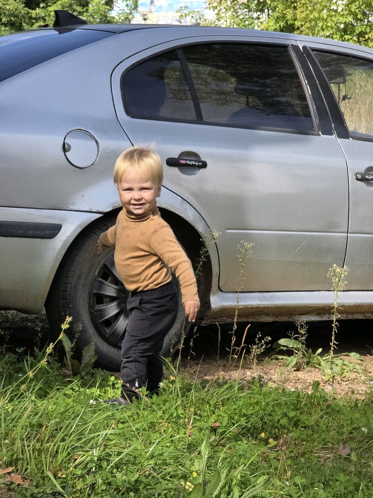 A young boy with blond hair is standing near a silver sedan in Chaikovsky, Russia. The boy is wearing a light brown turtleneck shirt with a pattern of small, cartoonish animals and black pants. He is looking towards the camera with a slight smile. The car is parked on a dirt surface, with green grass and foliage surrounding it. The car has a black shark fin antenna on the roof. A "TRD" decal is visible on the driver's side door handle. The scene appears to be outdoors on a sunny day.
