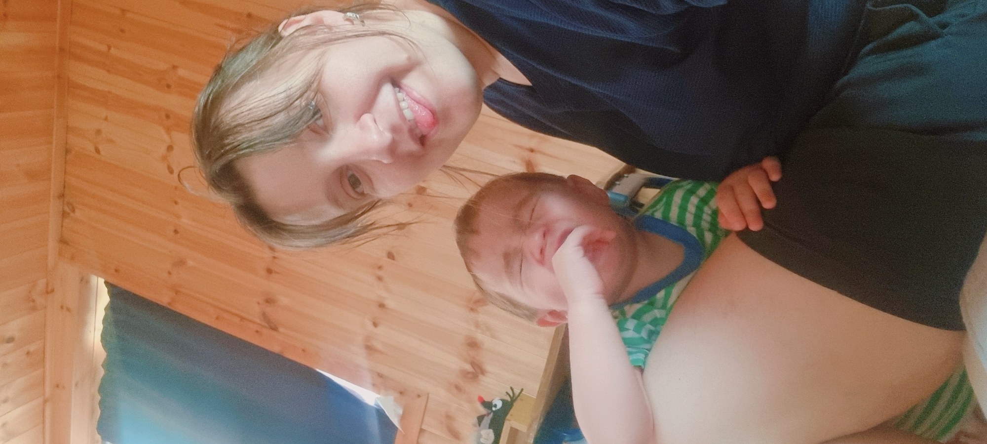 The image depicts a woman and a young child indoors, likely in a cabin or house with wooden walls and ceiling. The woman is smiling and sticking out her tongue towards the camera. The child appears upset, with a hand near their mouth, possibly crying. They are wearing a green and white striped shirt with blue trim. A stuffed mole toy is partially visible in the bottom left corner of the frame. The context does not provide information to determine the location within Dunnae-myeon, South Korea.