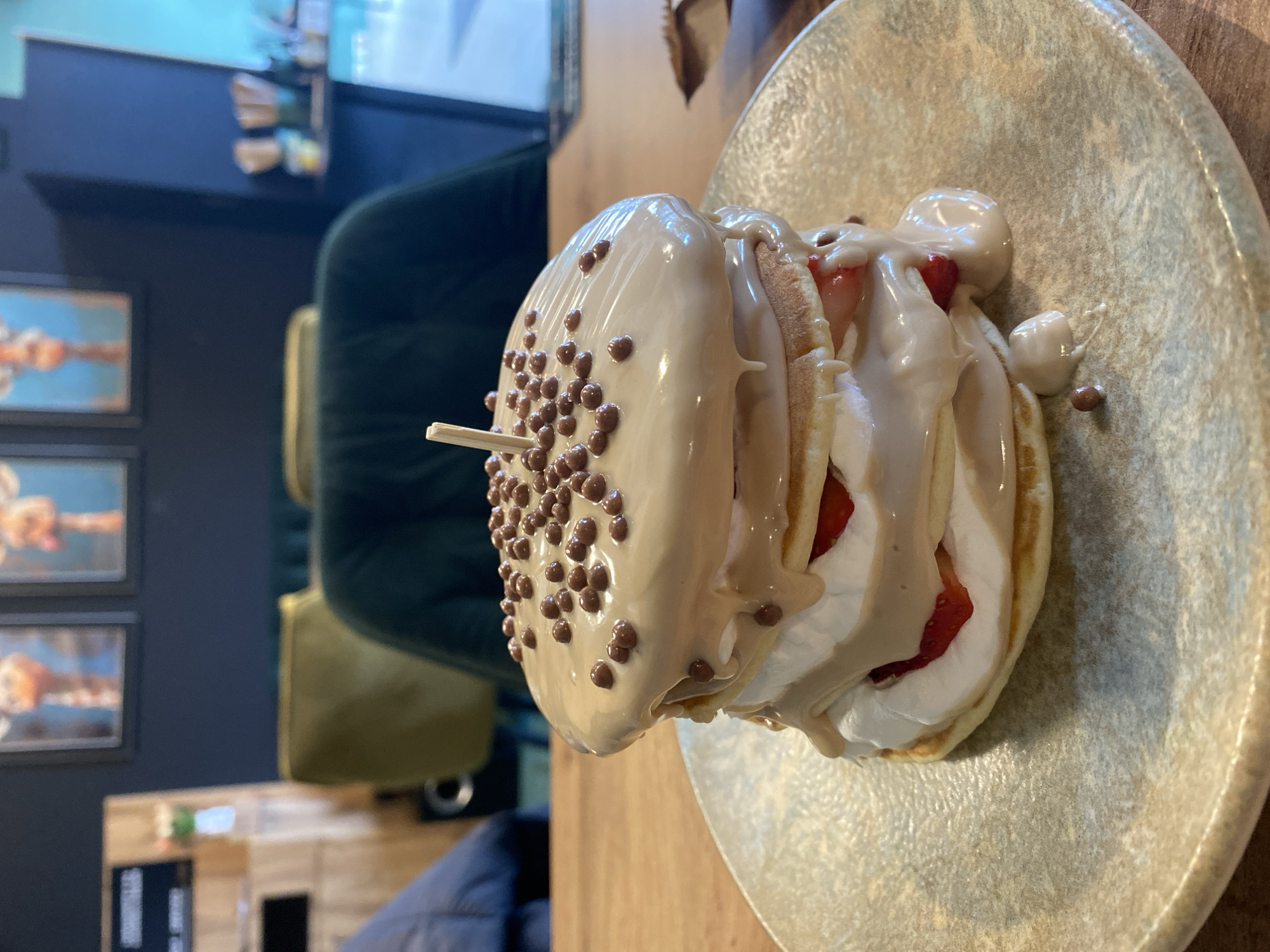 The image depicts a plate of stacked pancakes, seemingly in an indoor setting, possibly a restaurant or cafe. The pancakes are layered with cream and strawberries, and topped with a light brown sauce and small chocolate balls. A wooden toothpick pierces through the stack. The plate itself has a textured, light-colored surface, suggesting a ceramic or stoneware material. The table is made of wood. In the background, there is a dark green velvet chair, framed artworks on the wall, and a small piece of furniture. The setting conveys a casual dining atmosphere. The location in the city of Unknown, Unknown is impossible to determine from the image.