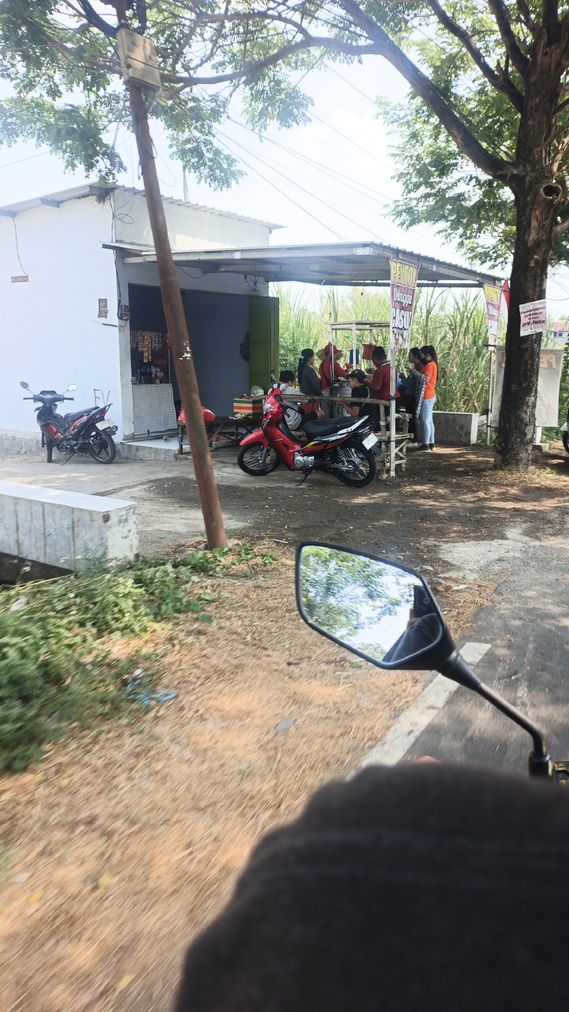 This is a candid, eye-level, exterior shot from the perspective of someone riding a motorcycle in Pohkecik, Indonesia. The scene captures a small, open-air food stall next to a white building. In the foreground, a motorcycle's rearview mirror and handlebars are visible, reflecting trees and the sky. The stall is shaded by a corrugated metal roof supported by wooden posts. Several people are gathered at the stall, some appearing to be customers and others staff, with food and drinks being served. Two motorcycles are parked near the stall, one red and one red and black. A tall wooden utility pole stands to the left of the stall. The ground is covered with dirt and dry grass, and lush green foliage is visible on the left side of the image. A concrete barrier runs along the left edge of the scene. In the background, sugarcane stalks are visible, indicating a rural or semi-rural setting. The overall impression is of a typical roadside scene in Indonesia, likely during daylight hours.