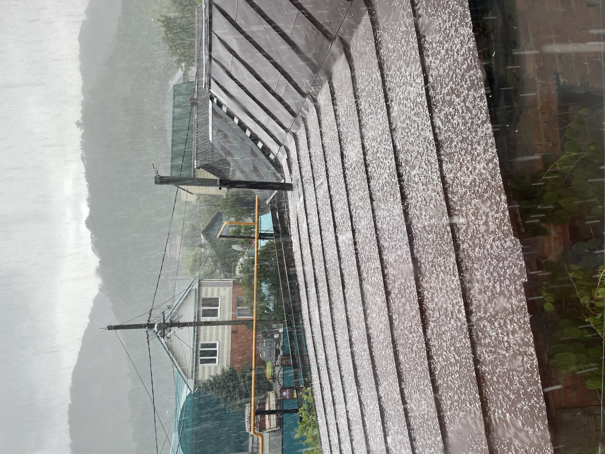 The image depicts a heavy downpour in what appears to be a residential area of Olginka, Russia. In the background, a misty mountain is visible, partially obscured by the rain and fog. Several houses and buildings, along with power lines and utility poles, are visible amidst the rain. A section of a building's roof is prominent in the foreground, with rainwater streaming down the corrugated surface. Some greenery is visible near a brick wall to the right of the roof. The overall impression is one of a heavy rainstorm in a mountainous or hilly settlement.