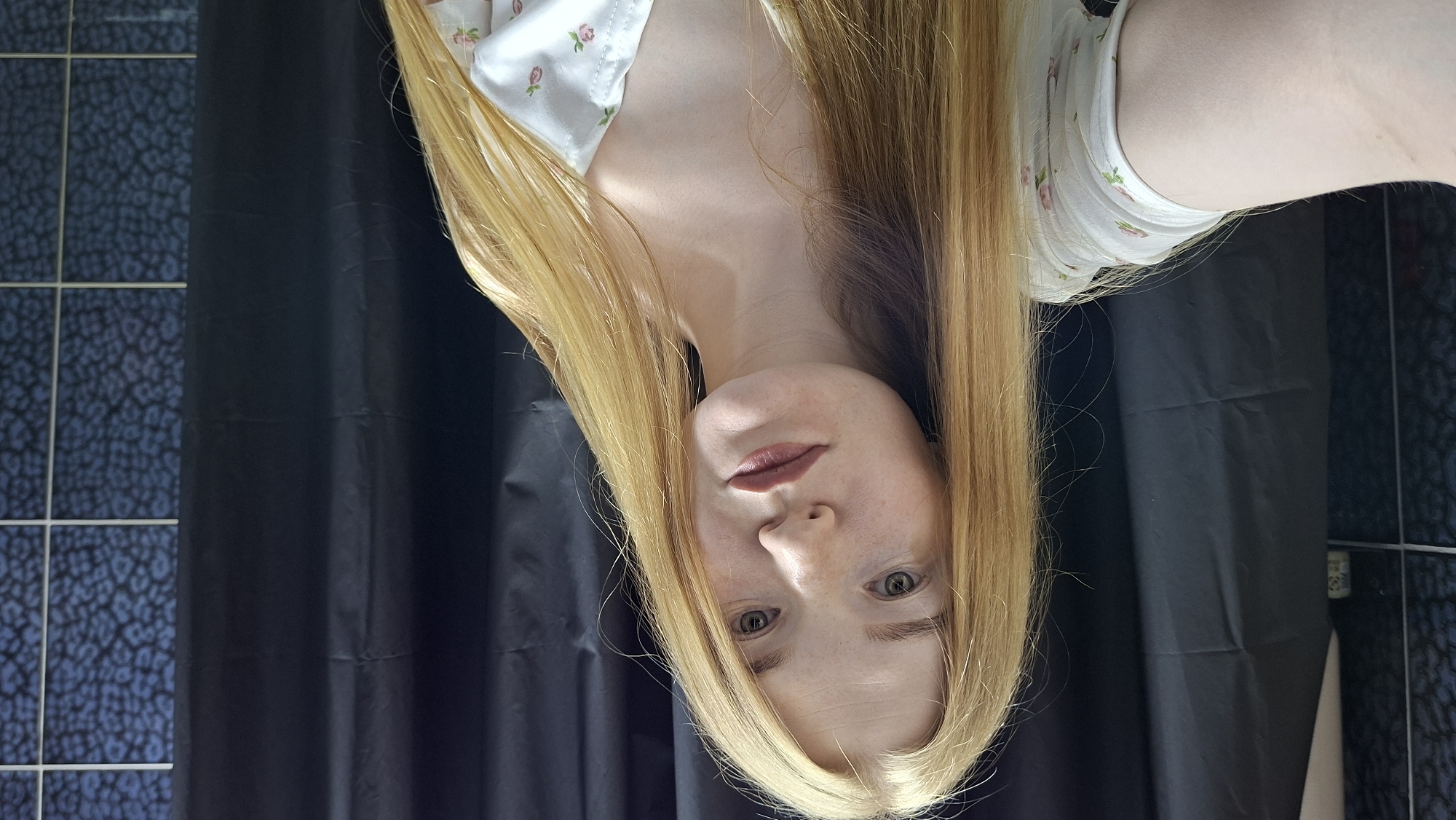 A young adult female is captured in a selfie photograph, appearing to be in a bathroom located in Chaikovsky, Russia. She possesses long, straight, light blonde hair that falls over her shoulders. Her complexion is fair with visible freckles, and she has light-colored eyes, likely green or hazel. A subtle, dark rose-colored lipstick is applied to her lips. She is wearing a light-colored top, possibly white or cream, adorned with a delicate pattern of small pink roses and green leaves, featuring a V-neckline. The background on the left side of the frame reveals a dark blue or black shower curtain, partially obscuring a dark tiled wall. A small, light-colored object rests on a shelf or ledge in this area. On the right side of the frame, a segment of the dark shower curtain is visible, adjacent to a blue tiled wall featuring a mottled or leopard-like pattern, delineated by white grout lines.