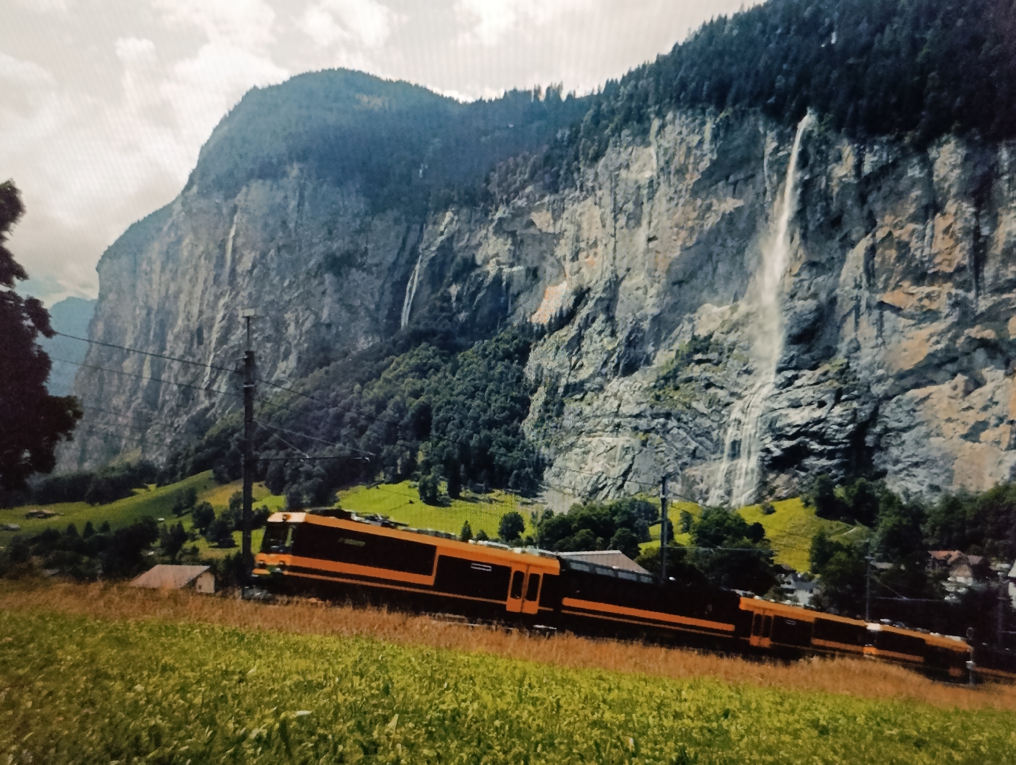 The image depicts a scenic mountain landscape with waterfalls cascading down a rocky cliff face. A train with orange and black carriages is seen traversing a grassy hillside in the foreground. The scene appears to be a natural environment, possibly a tourist attraction, with lush green vegetation and a cloudy sky. There are no discernible elements that would suggest the location is the city of Dubăsari town council, Moldova. The image focuses on the natural beauty of the mountains and the train as a mode of transport within this environment.