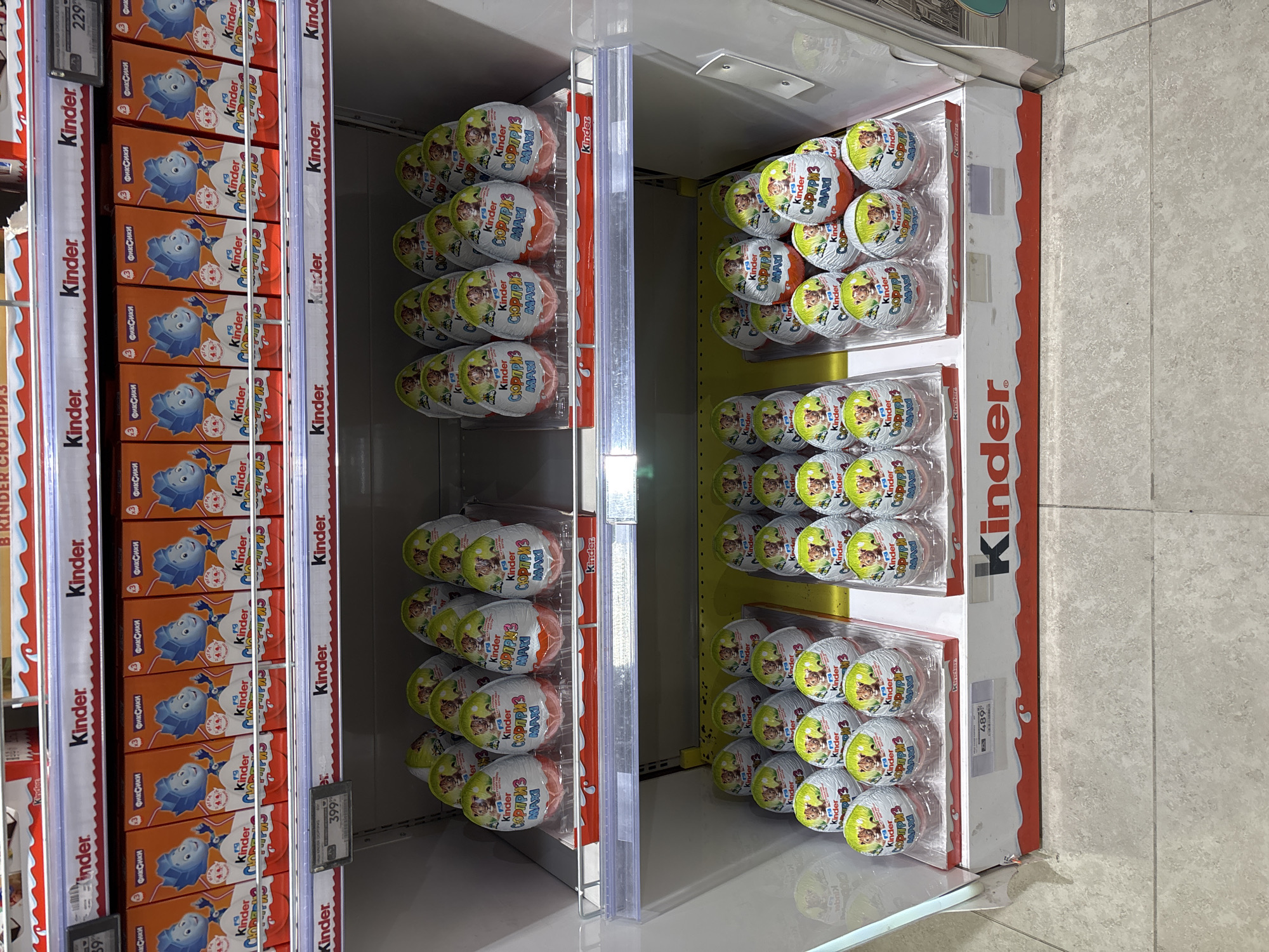 The image captures a retail store display filled with Kinder chocolate products. The scene is an aisle or a dedicated section within what appears to be a supermarket or convenience store, suggested by the shelving and product arrangement. No people are visible, but the setup indicates products are ready for purchase.
On the left side of the display, multiple rows of orange-colored Kinder chocolate bars or boxes are stacked vertically. These packages prominently feature characters from the Russian animated series "Фиксики" (Fixiki), indicating a themed product line. The text "Kinder" is also visible in English and Cyrillic script.
On the right side, several rows of Kinder Surprise Maxi eggs are neatly arranged on angled shelves. These eggs are individually wrapped in white, green, and yellow foil, with the words "Kinder" and "СЮРПРИЗ MAXI" (SURPRISE MAXI) clearly printed on them. These appear to be larger versions of the classic Kinder Surprise.
The products are organized within clear plastic shelf dividers. Price tags are visible at different points on the shelves, showing numbers such as "229", "399", and "489", likely representing prices in rubles, which aligns with the "Russia" location context. The overall display is well-lit and tidy, typical of a retail environment.