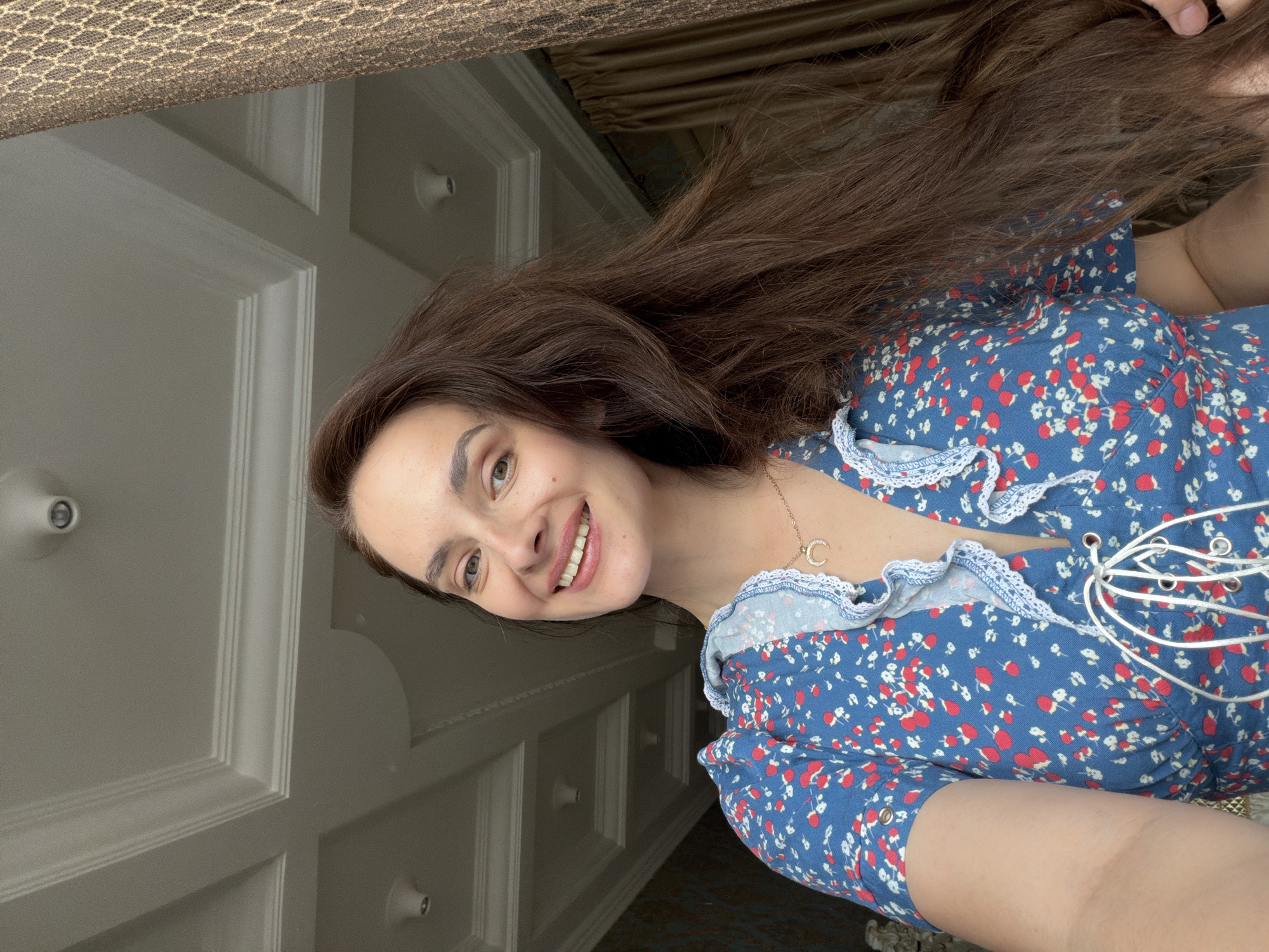 This is a portrait of a young woman, likely taken indoors. She has long, dark brown hair that cascades around her. She is wearing a blue dress with a floral pattern of red and white flowers. The dress features a ruffled neckline and a lace-up detail. A delicate gold necklace with a crescent moon pendant rests on her collarbone. She is smiling warmly at the camera, her eyes conveying happiness.
The background appears to be a room with light-colored paneled walls, suggesting a home environment. The lighting suggests it could be daytime. The overall mood of the image is cheerful and relaxed.
