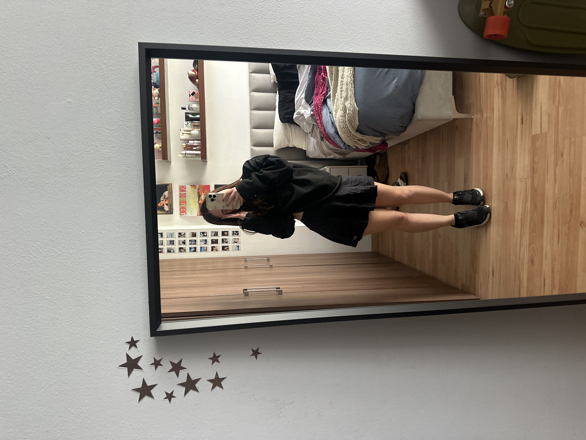 A person stands in a room, visible in a mirror hanging on a white wall decorated with star decals. The individual holds a smartphone, obscuring part of their face. They wear a black top and shorts, with dark athletic shoes. Reflected in the mirror is a room with wood flooring and a light-gray upholstered headboard. A bed is partially visible, covered with various layers of blankets and bedding in shades of gray, white, and red. To the left of the mirror reflection is a shelf of personal care products and a collage of photos. A wood-paneled closet with horizontal silver handles is also reflected. A skateboard is partially visible in the upper right corner of the mirror’s reflection. The location is Huixquilucan de Degollado, Mexico.