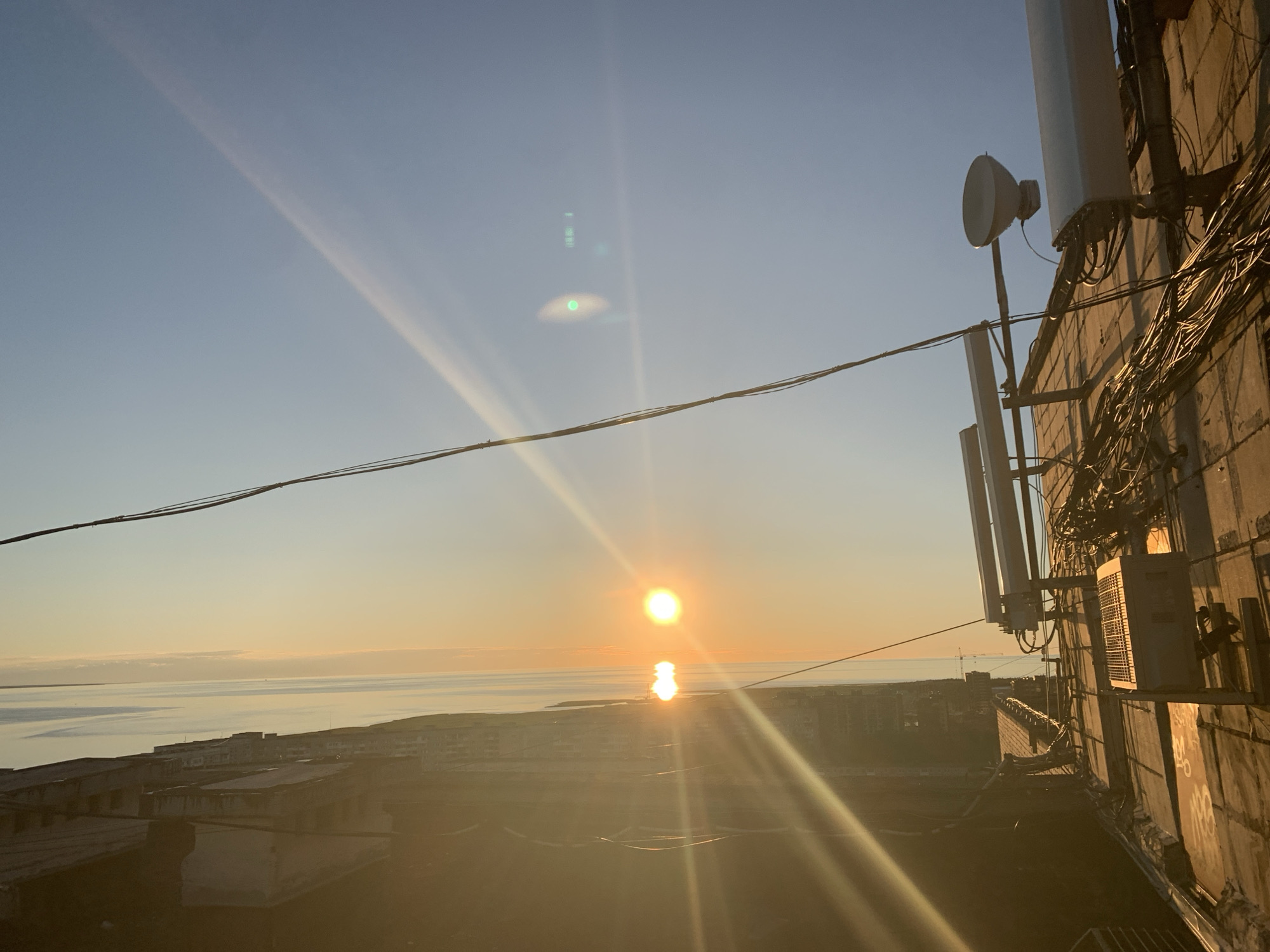 The image depicts a sunset over a body of water, viewed from a high vantage point in the city of Severodvinsk, Russia. The foreground is dominated by the side of a building with multiple telecommunication antennas and a complex network of wires attached to its facade. The building's wall appears to be made of concrete blocks. In the midground, a low-rise urban landscape of buildings is visible, with flat roofs and some appearing to be in a state of disrepair. The background features a calm body of water, likely the White Sea, meeting the horizon where the setting sun casts a golden glow. The sky transitions from a clear blue at the top to a warm orange and yellow hue near the horizon, with visible sun flares and lens artifacts. There are no people or animals present in the image. The overall scene conveys a sense of an industrial or coastal city at dusk.