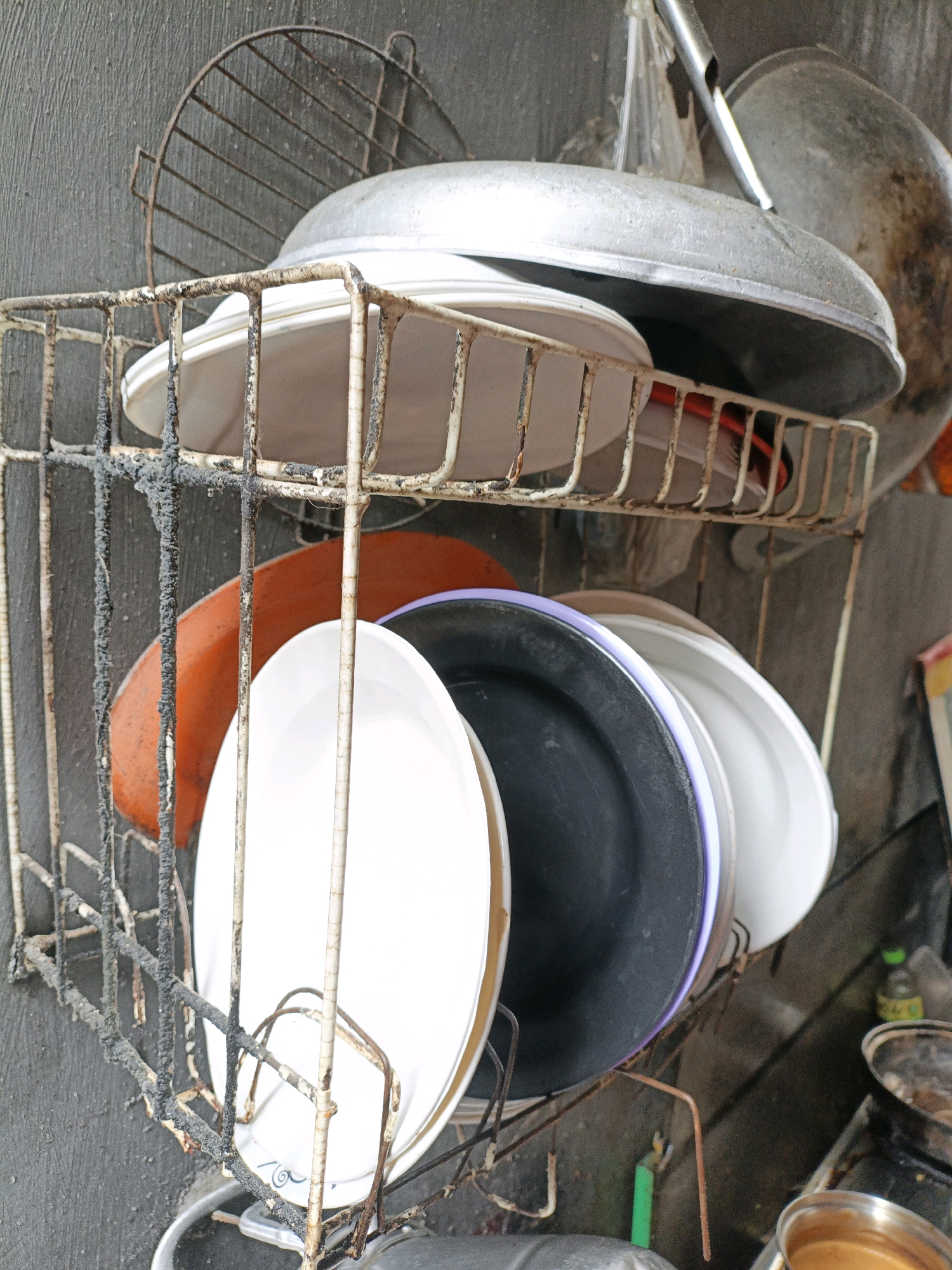 An outdoor dish rack holds an assortment of plates and a large metal wok. The dish rack is attached to a gray, textured wall, and appears to be made of rusted metal with significant buildup of grime and possibly carbonized residue. In the foreground, several plates are stacked, including a large white plate with a circular design near the rim, a black plate with a purple edge, and an orange plate. Above these, a large, shallow metal wok is positioned on top of the rack, its underside showing signs of heavy use and discoloration. The scene is set in an outdoor environment, possibly a backyard or alleyway, suggesting a utilitarian or rustic setting. The image was taken in West Jakarta, Indonesia.