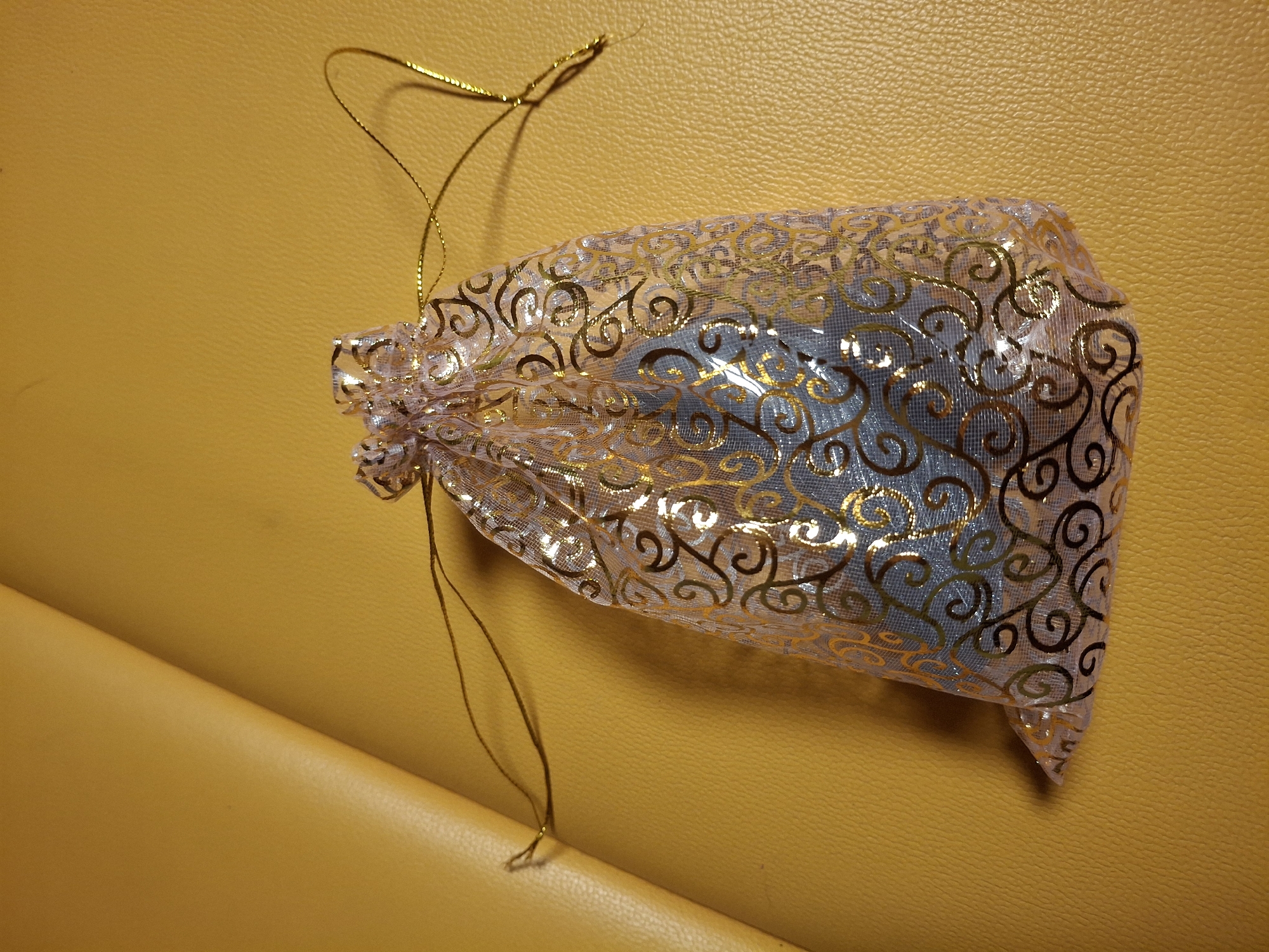 This is a close-up, eye-level shot of a small, translucent drawstring bag with a golden scroll pattern. The bag appears to be made of a fine mesh material and is filled with a light blue object, possibly jewelry or a small gift. The bag is cinched closed at the top with a thin, golden cord that extends upwards and to the left, casting a subtle shadow. The bag is placed on a textured, yellow surface, which could be leather or a similar material. The lighting suggests an indoor setting, likely with artificial light, creating soft shadows and highlighting the sheen of the bag and its pattern. No people are visible, and the focus is entirely on the object and its immediate surroundings. The image lacks any specific location cues beyond the provided context of Ogre, Latvia, and does not show any text. The overall impression is one of a small, decorative item, possibly prepared for gifting.