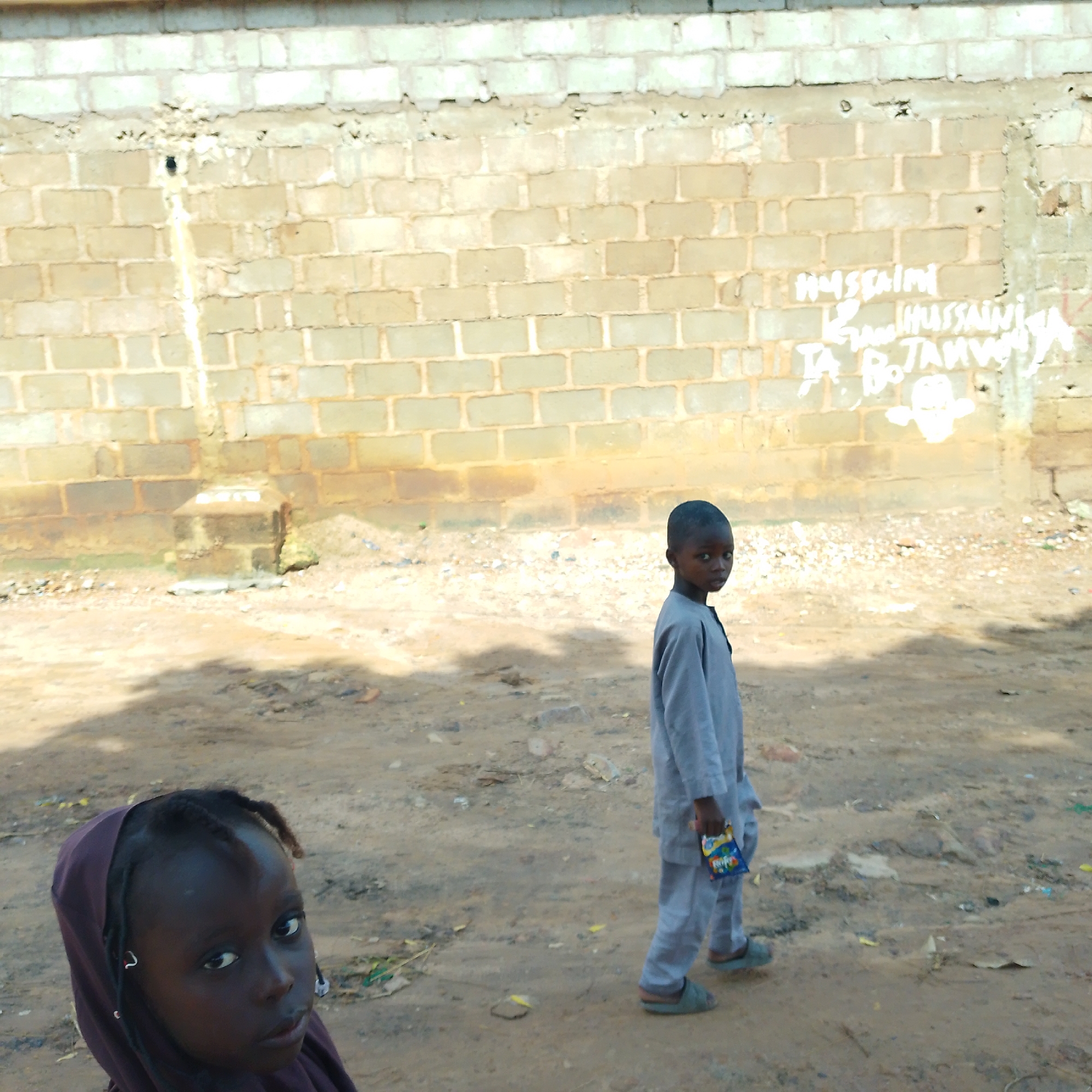 In Pantame, Nigeria, two children are pictured outdoors in front of a brick wall. The child on the left, a young girl wearing a dark purple head covering, is in the foreground and is looking up and to the left. The child on the right, a boy wearing a light grey outfit and sandals, is walking to the left and looking at the camera. He is holding a colorful packet in his right hand. The ground is dirt with scattered debris. The brick wall behind them has writing on it, including "HUSSAINI" and "TA. BO TANWO".