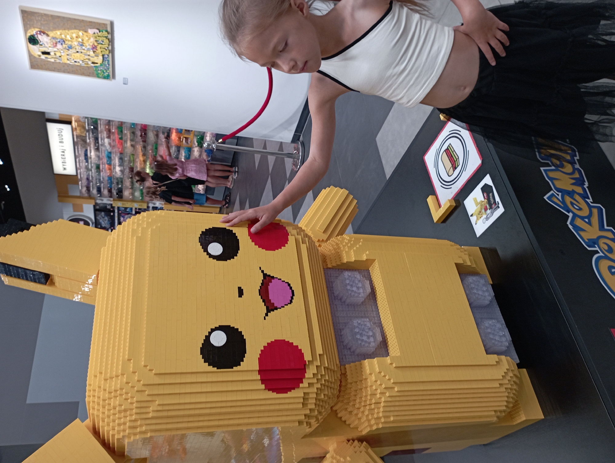 A young girl with blonde hair, wearing a white tank top and a black tulle skirt, stands to the right of a large, yellow LEGO statue of the character Pikachu. She is touching Pikachu's left cheek with her right hand. In the background, a store displays shelves of colorful LEGO bricks. Above the shelves, a sign reads "WYBIERAJ I BUDUJ" (Choose and Build). A framed mosaic artwork, resembling Gustav Klimt's "The Kiss," hangs on the white wall to the right. A red velvet rope barrier is visible, separating the area. The scene appears to be indoors, possibly in a retail or exhibition space. The LEGO Pikachu is a prominent subject, with its characteristic large ears, black eyes with white pupils, red circular cheeks, and open mouth with a pink tongue. The environment is well-lit, suggesting artificial lighting. A small sign with a sandwich icon and a Pikachu sticker are on a black surface in front of the girl and the LEGO statue. The lower part of the image shows a black surface with a portion of a blue and yellow logo that reads "Pokém..."