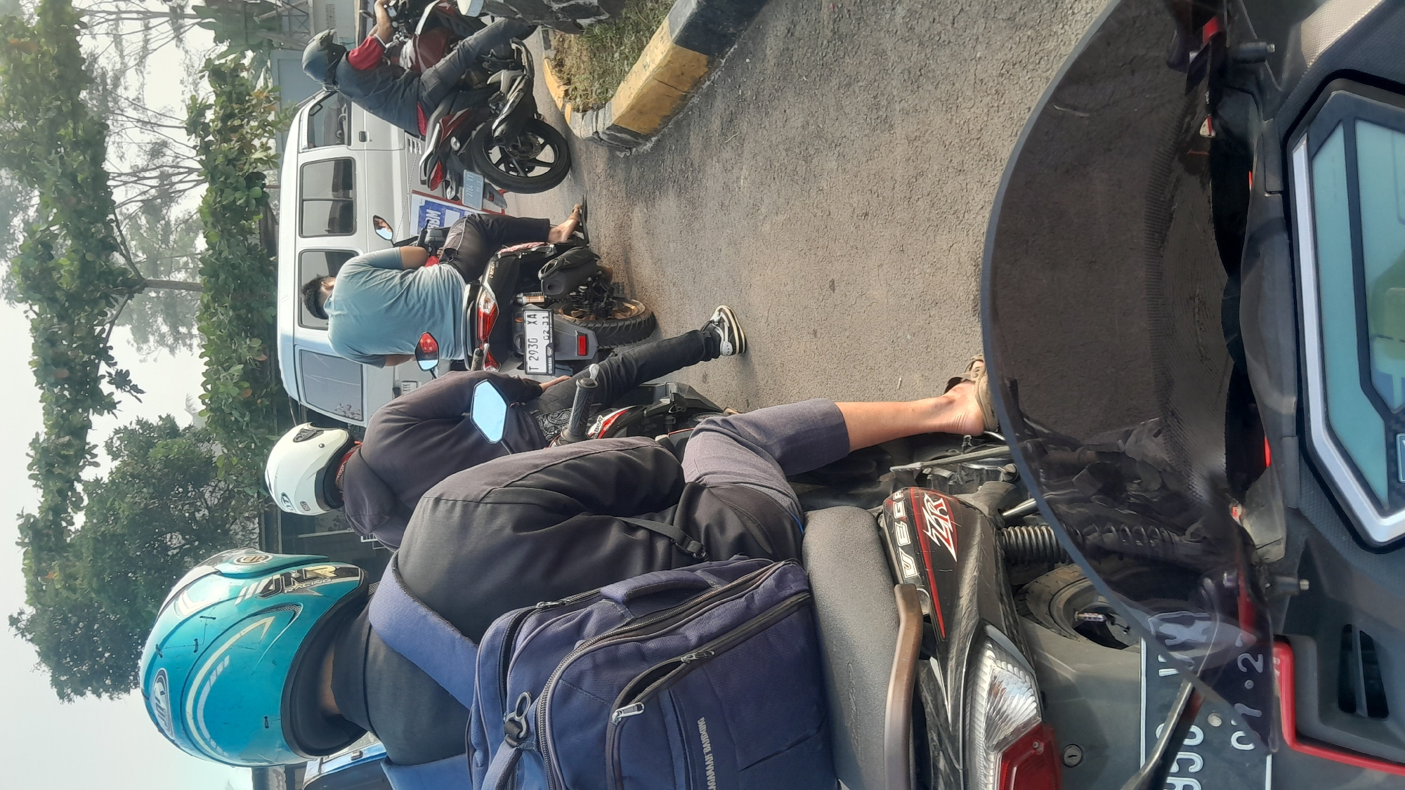 The image shows a traffic scene in Subang, Indonesia, focusing on motorcycles lined up on a road. In the foreground, the front of a motorcycle with a dark, reflective windshield and a digital display is visible. Behind this, another motorcycle rider wearing a black jacket and a blue backpack on their back is seated. To their right, a person in a grey hoodie, wearing a white helmet, is on a motorcycle. Further ahead, a person in a light blue t-shirt and dark pants is seated on a motorcycle. On the right side of the image, a person in a red shirt and black vest is riding a motorcycle. A white van is visible in the background, along with trees and vegetation. The road has a yellow and black curb on the right side. The scene appears to be a traffic jam or a waiting area.