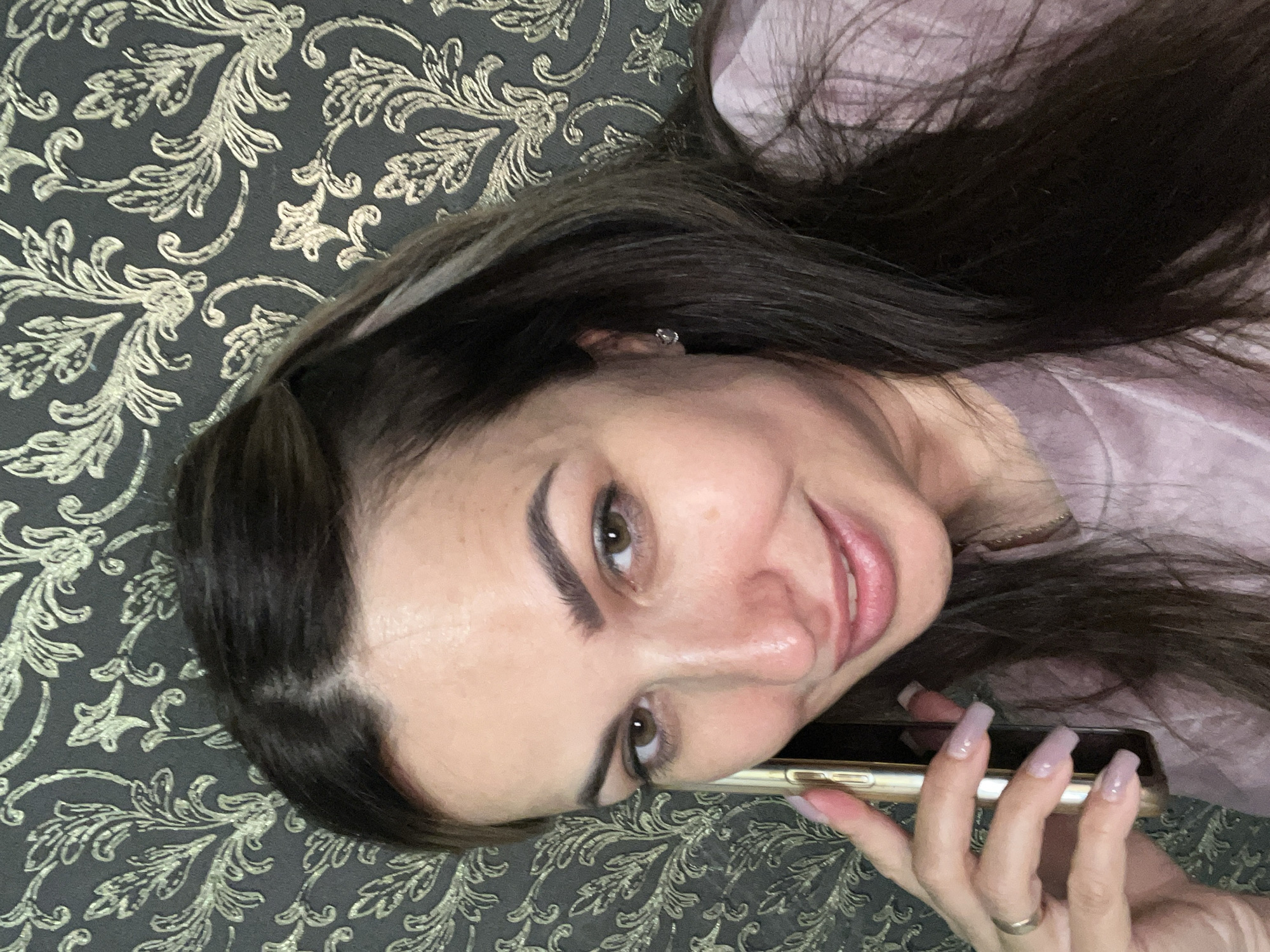 A medium shot of a woman with dark hair and brown eyes is taken from a slightly high angle. She is holding a gold-colored smartphone to her right ear with her left hand and smiling at the camera. Her left hand has a gold ring on the ring finger and long, light purple painted nails. She is wearing a light pink t-shirt. The background is a dark gray wallpaper with a gold ornate floral pattern. The scene appears to be indoors, possibly in a home, with no indication of a specific city or location.