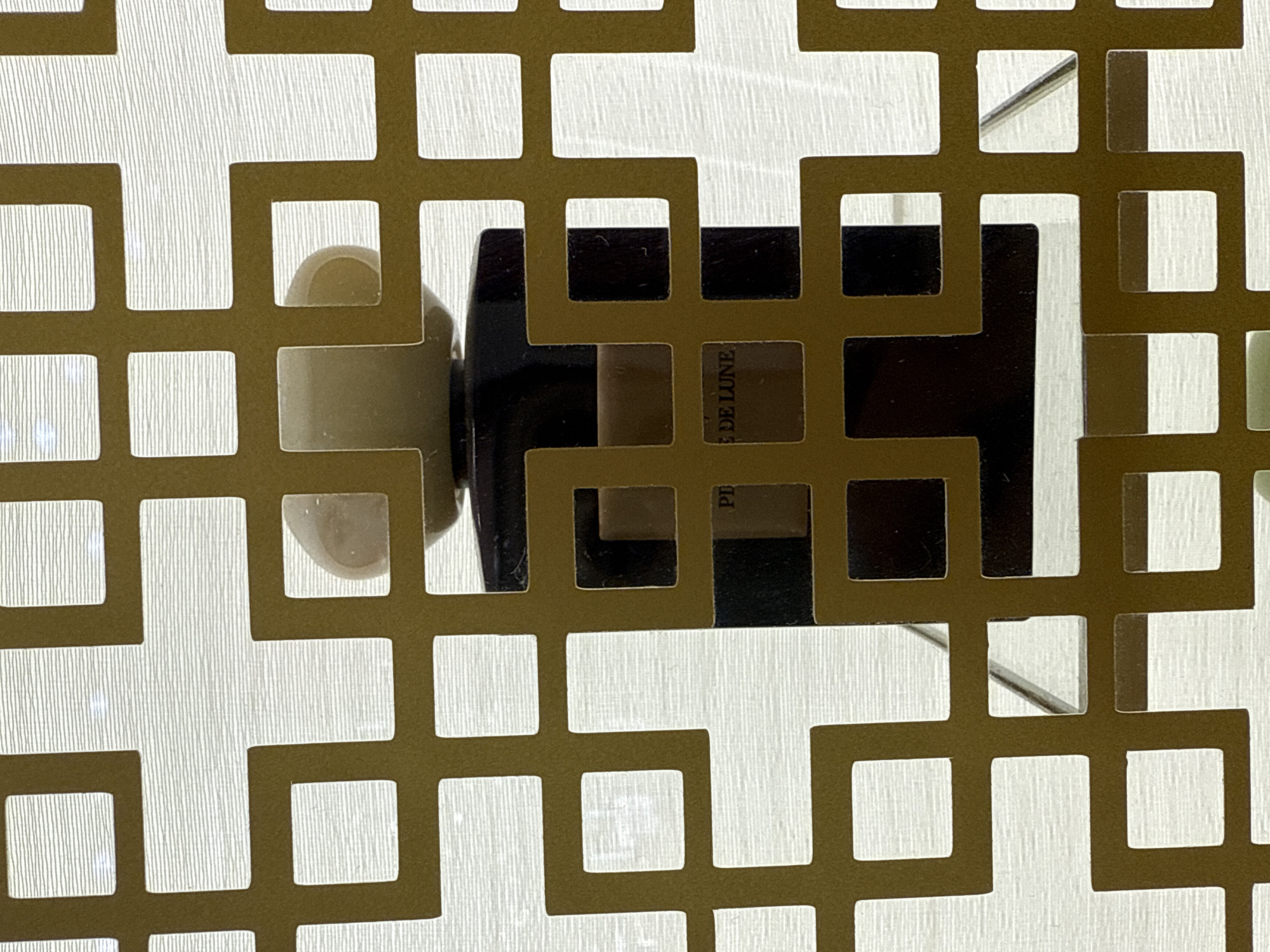 A perfume bottle with a black base and a light-colored cap is visible through a decorative gold lattice screen. The bottle has text that reads "PI DE LUNE" on its label. The background appears to be a light-colored textured surface, possibly a wall or fabric. The scene is indoors, and no specific location in Astana, Kazakhstan, can be determined from the image alone. There are no people or actions depicted.