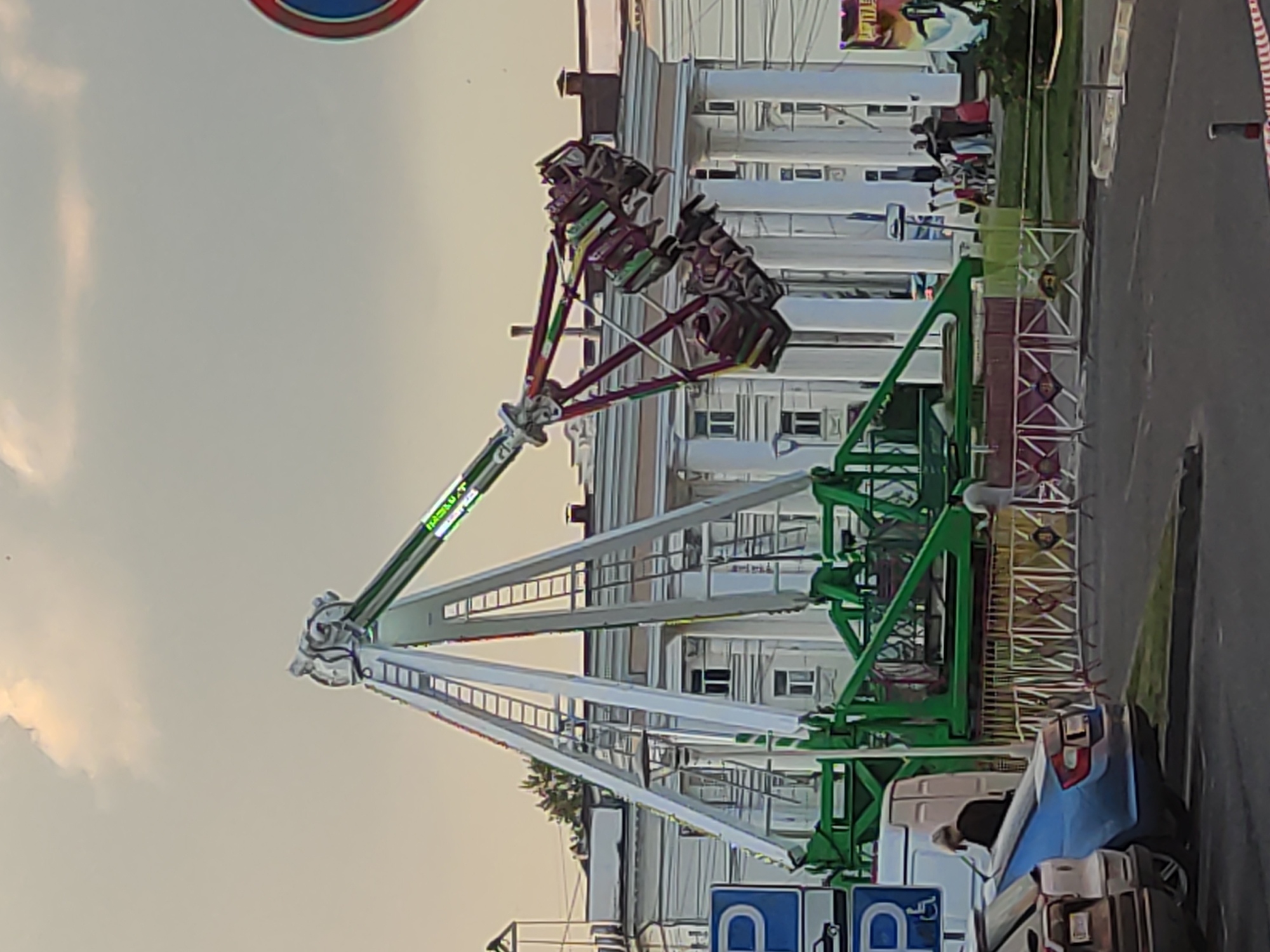 The image shows a large amusement park ride, likely a pendulum ride, in motion. The ride has a central tower structure with a long arm extending outwards, from which suspended seats are swinging. The seats appear to be occupied by people. The ride is located in an outdoor setting, with a building in the background that features a classical architectural style with columns. There are also cars parked in the foreground and a road. People are visible near the ride and on the sidewalk. The sky is overcast with some lighter clouds. The image appears to be taken during the daytime. Based on available data, the image is located in Zhirnosovo, Russia.