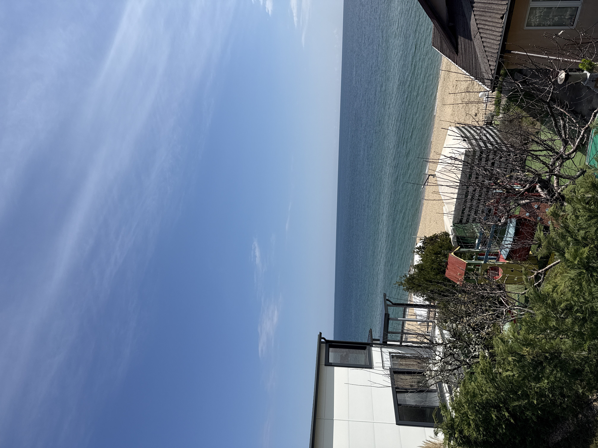 The image depicts a beach scene in Varna, Bulgaria. The right side of the image features a sandy beach, bordering the sea. Adjacent to the beach is residential development, including a white building with dark-framed windows and a tan building with a dark roof. The foreground vegetation consists of trees and shrubbery, partially obscuring a playground structure with a red roof. The left side of the image shows a clear blue sky with wispy clouds.