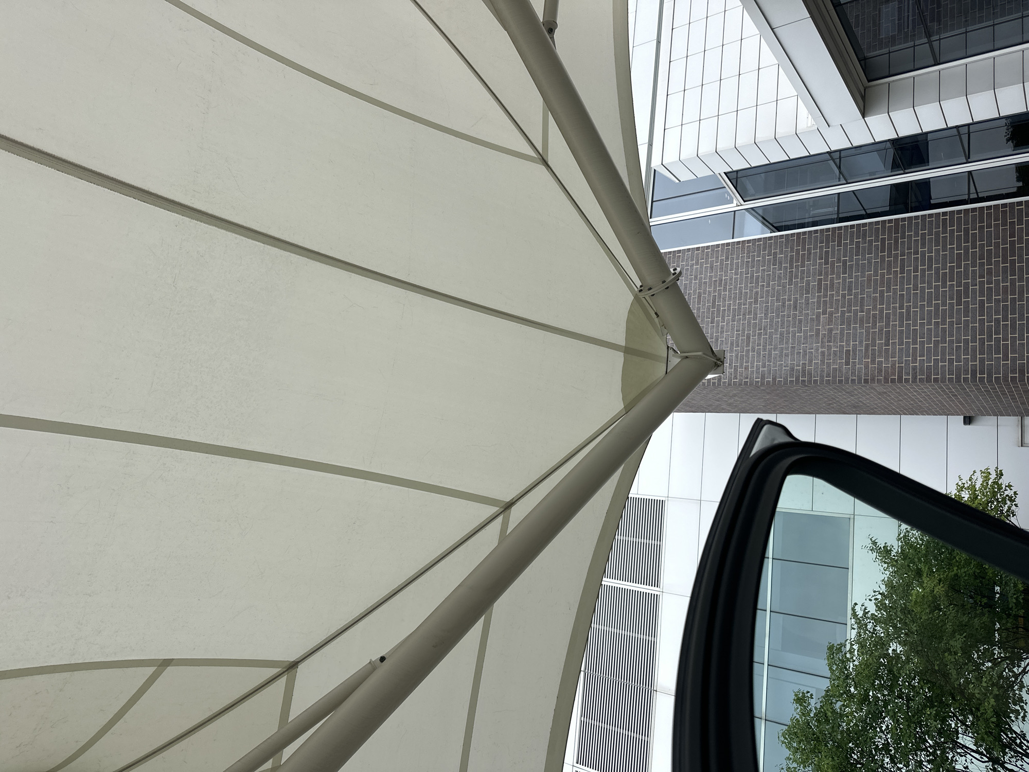 A low-angle shot captures the underside of a light-colored fabric awning supported by a metal frame. The awning is segmented into panels by darker seams and extends over a modern building. To the left, the open door of a vehicle reveals a green tree and a building with horizontal vents and large windows. The building features a facade of white panels and dark brick.