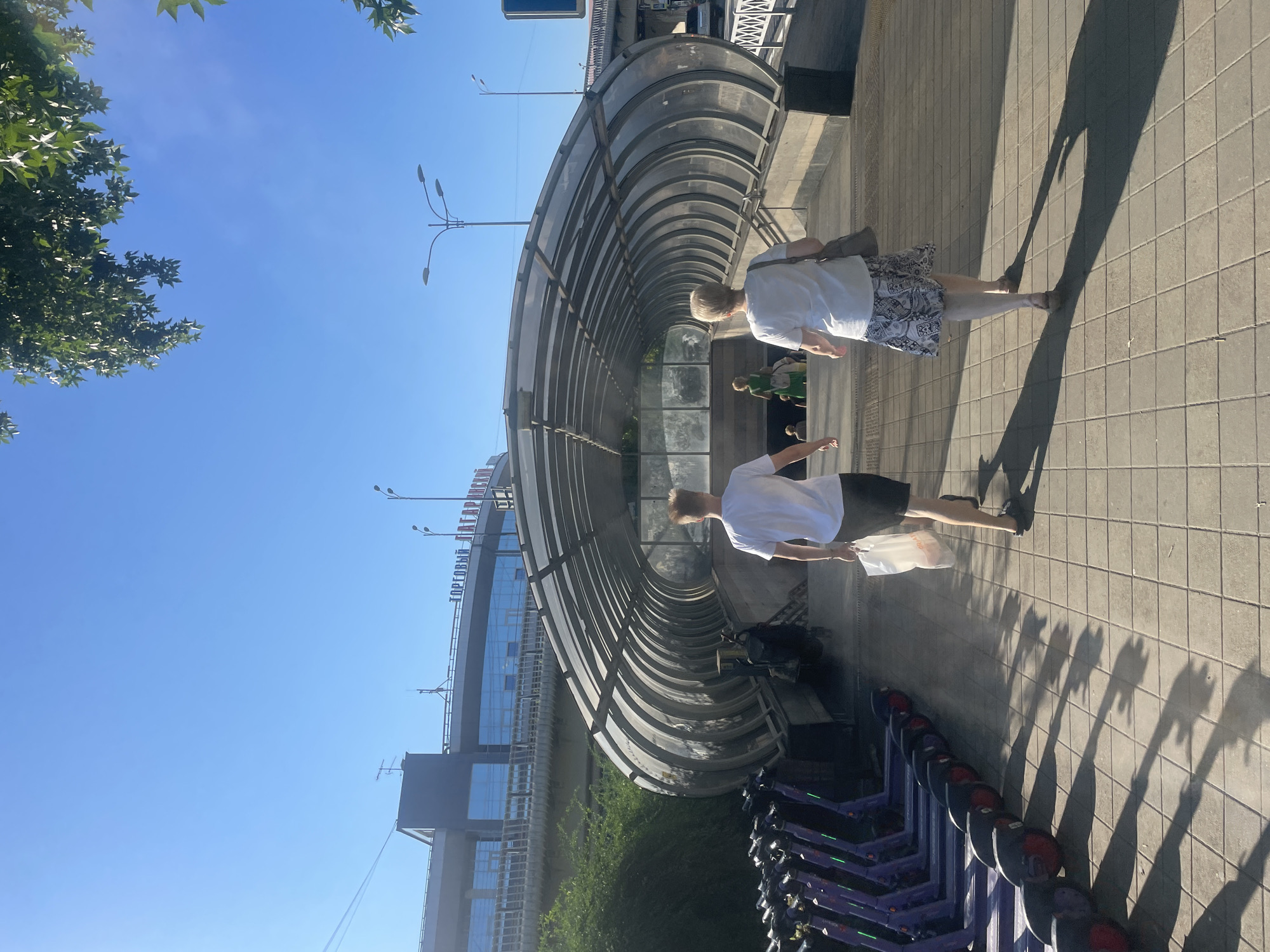 The image depicts an outdoor scene on a sunny day, featuring a pedestrian tunnel with a glass and metal arched structure leading downwards. Two adults, a man and a woman, are walking away from the camera towards the tunnel entrance. The man, wearing a white t-shirt and black shorts, carries a white plastic bag. The woman, in a white t-shirt and a patterned skirt, carries a grey bag over her shoulder. Further into the tunnel, two more people, one in a green dress, are visible. To the left of the path leading to the tunnel, a row of parked purple electric scooters is lined up. In the background, a modern building with large windows and signs reading "ТОРГОВЫЙ ЦЕНТР" (Shopping Center) is visible, along with streetlights and trees. The ground is paved with square tiles, casting long shadows from the scooters and people. The sky is clear and blue. This scene is likely located in Sochi, Russia, given the context provided.