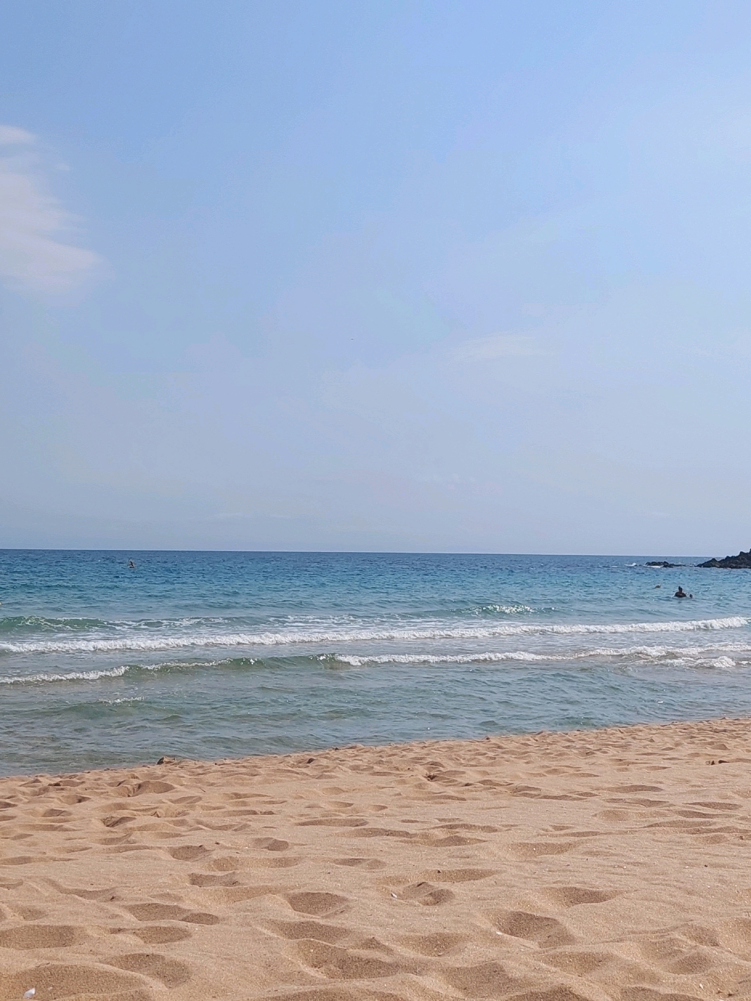The image depicts a serene beach scene, likely from Sozopol, Bulgaria, given the context. In the foreground, soft, light brown sand covers the majority of the frame, showing subtle undulations and scattered indentations, possibly from footprints. The sand transitions into the shallow waters of the sea, where gentle, translucent waves with white foamy crests are breaking and receding.
Further out, the sea deepens to a rich blue, stretching towards the horizon where it meets a pale, hazy sky. The horizon line is clear and distinct. To the far right of the image, a dark, rocky outcropping juts into the sea.
In the mid-distance of the water, two individuals are visible. One appears to be swimming or wading, with their body mostly submerged. Another person is further out in the water, closer to the rocky outcrop, also appearing to be enjoying the sea. The overall impression is one of peacefulness and a typical summer day at the coast.