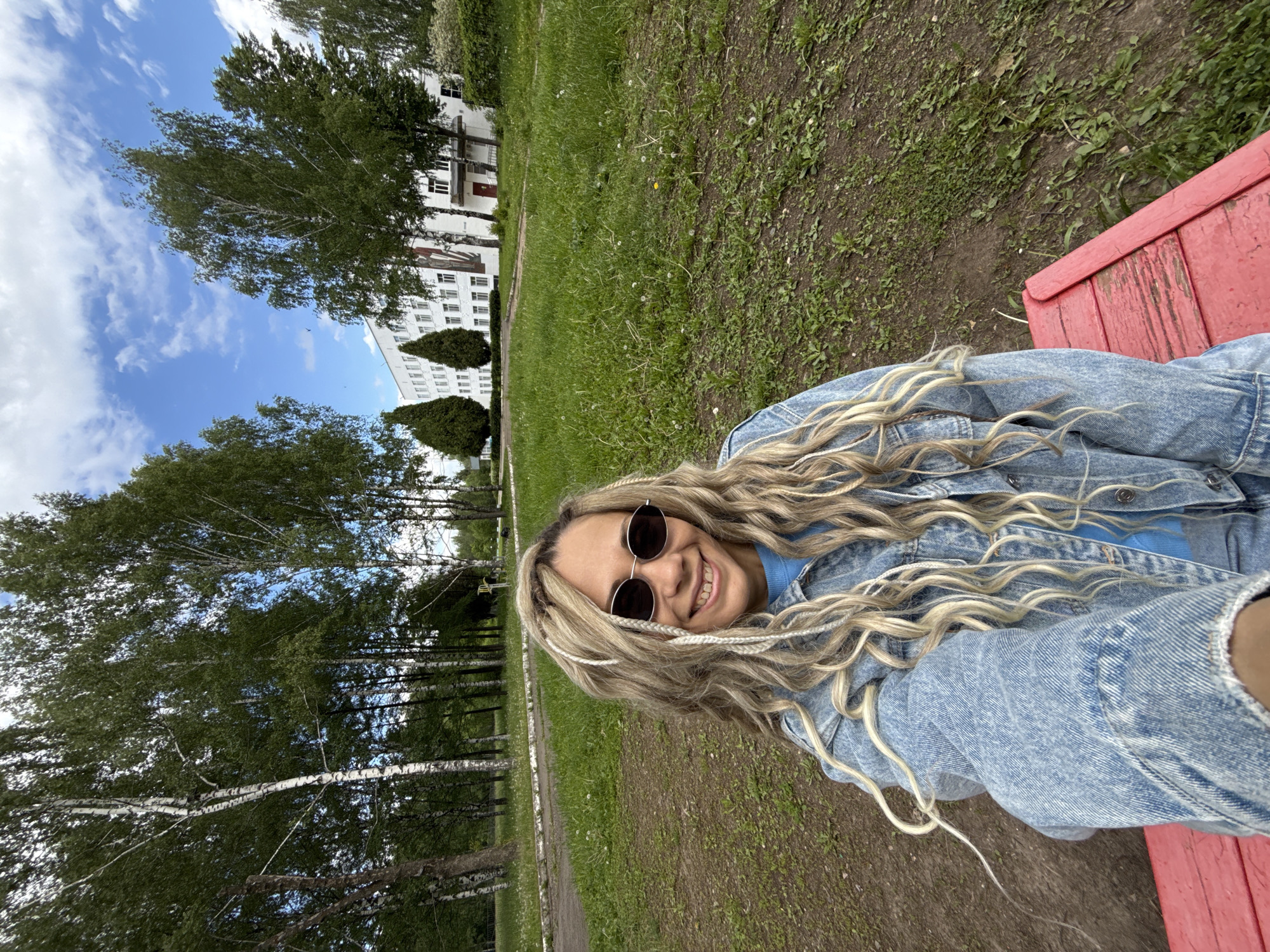 The image shows a woman sitting on a red bench in Шебунино, Russia. She is wearing a blue denim jacket, a blue shirt, and round sunglasses. She has blonde braids. Behind her is a grassy area with trees and a white building. The sky is visible with some clouds.