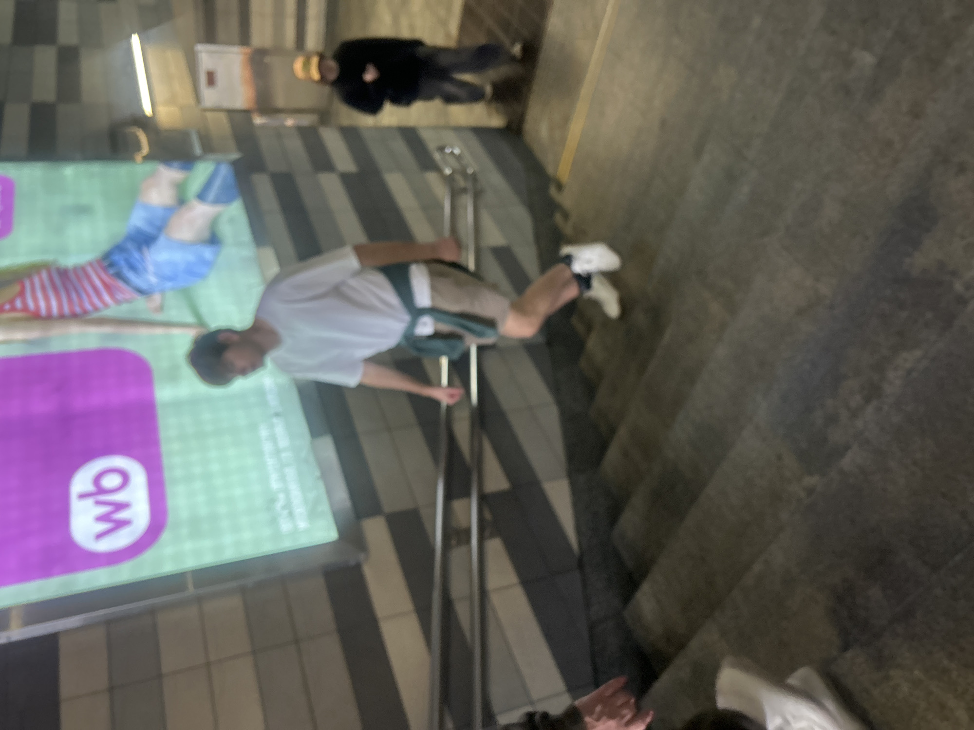 The image shows a blurred view of people on a set of outdoor stairs. A young man in a white t-shirt and beige shorts, with a green jacket tied around his waist, is descending the stairs. He is wearing white sneakers. Behind him and to the right, another person in a dark hoodie and a yellow cap is also walking down the stairs. In the background, a large advertisement billboard with the text "wb" is visible. The stairs appear to be made of concrete. The overall lighting suggests it is either evening or indoors with artificial lighting. The location is not identifiable as Moscow, Russia, based on the visual information provided.