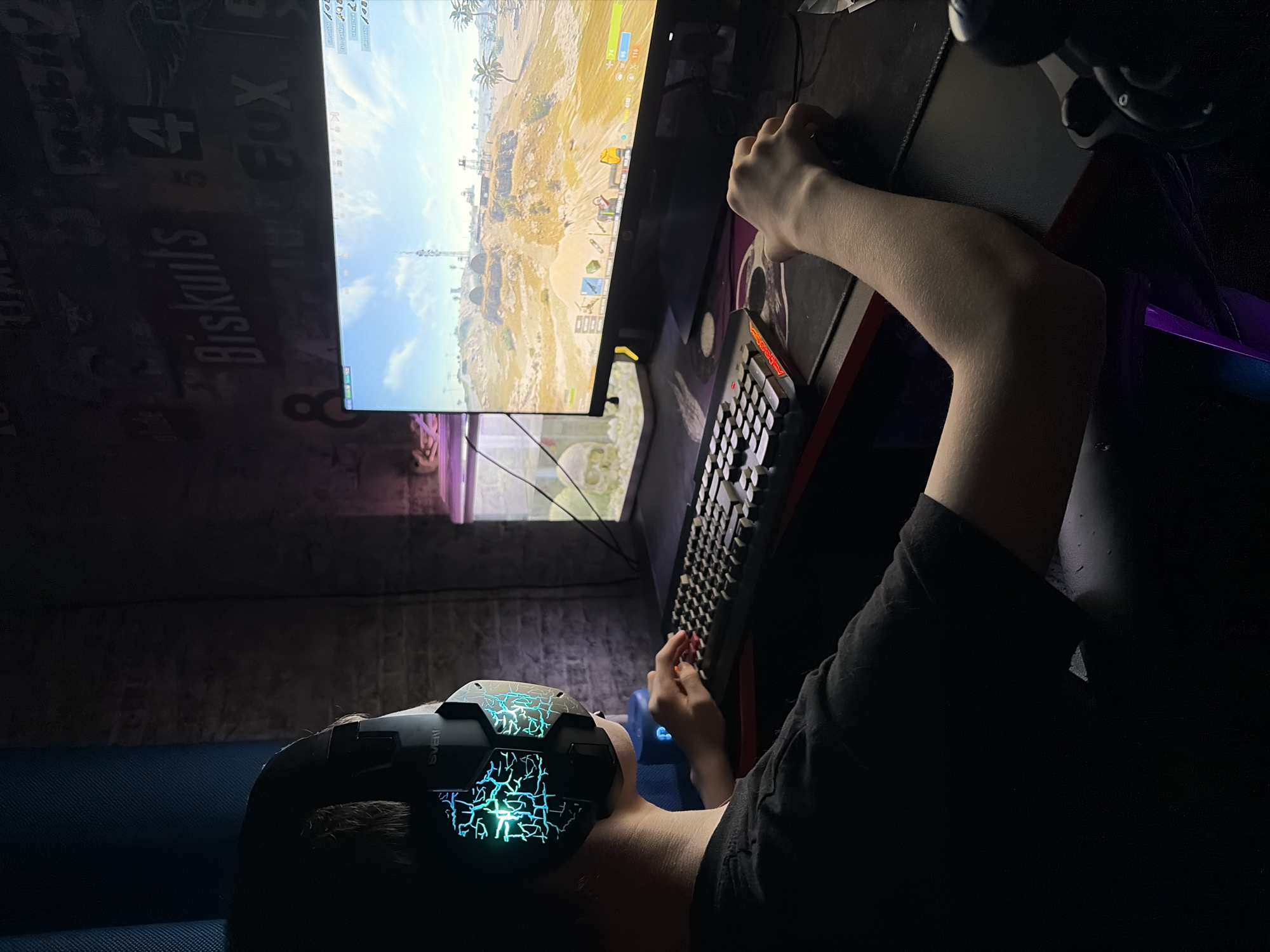An individual is depicted seated at a desk, engaged in playing a video game on a computer monitor, within a dimly lit room in the city of Chaikovsky, Russia. The person, appearing to be a male youth or young adult, is shown from a right-side profile perspective, wearing a black t-shirt and black gaming headphones. The headphones feature prominent illuminated earcups with a blue and green crackled design, and the brand "SVEN" is visible on the left earcup. The person's left hand rests on a black gaming keyboard, which displays red backlighting on some keys, while their right hand operates a black computer mouse on a dark gaming mousepad.
The computer monitor displays an active video game, identified by its user interface elements and a desert-like landscape featuring structures, a tall radio tower, and palm trees under a partly cloudy sky. To the immediate left of the monitor, a small blue cylindrical device, possibly a speaker, is visible. Further to the left, a glass tank, likely an aquarium or terrarium, contains a skull decoration and is illuminated by a subtle purple light emanating from its vicinity.
The wall behind the desk is dark-toned and textured, possibly brick or concrete, adorned with various white stenciled or painted words and numbers, including "Biskuits," "4," "8," and "Fox." A section of blue fabric or curtain is visible in the upper left corner of the frame. The overall environment suggests a dedicated personal gaming setup.