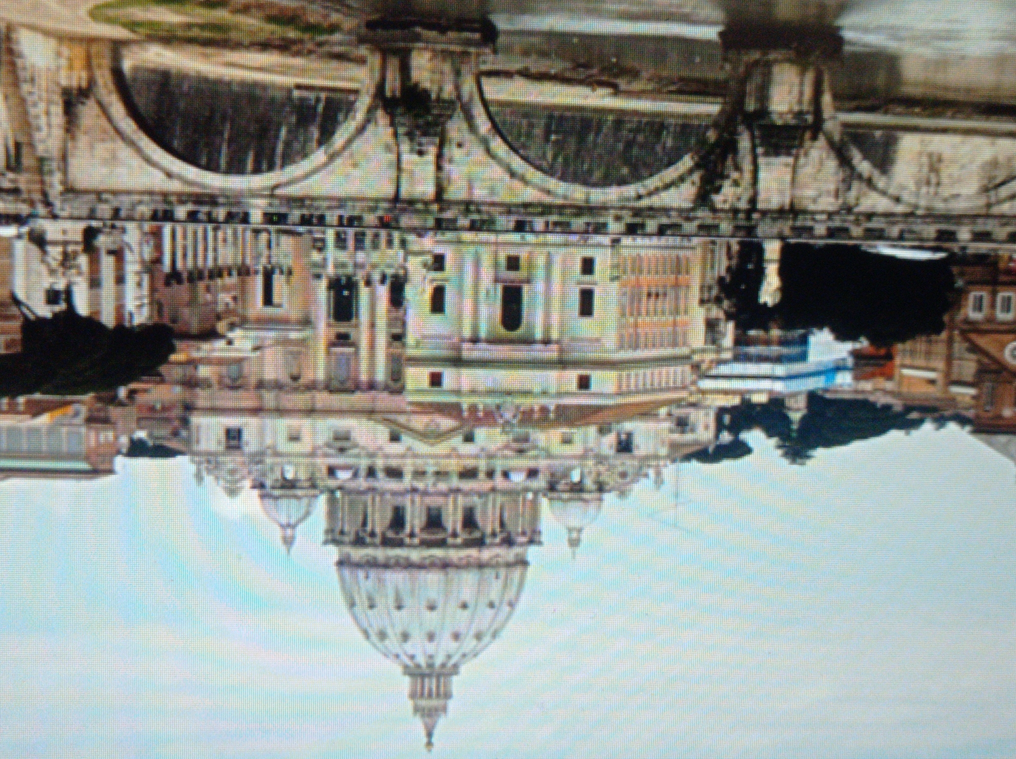 The image shows a large domed building, likely a cathedral or basilica, with classical architectural elements such as columns and arches. In the foreground, there is a stone bridge with multiple arches spanning a body of water. The bridge is made of weathered stone, and there are some trees and greenery visible on its surface. The sky is overcast. The description does not mention Mukachevo, Ukraine, as this is not depicted in the image.