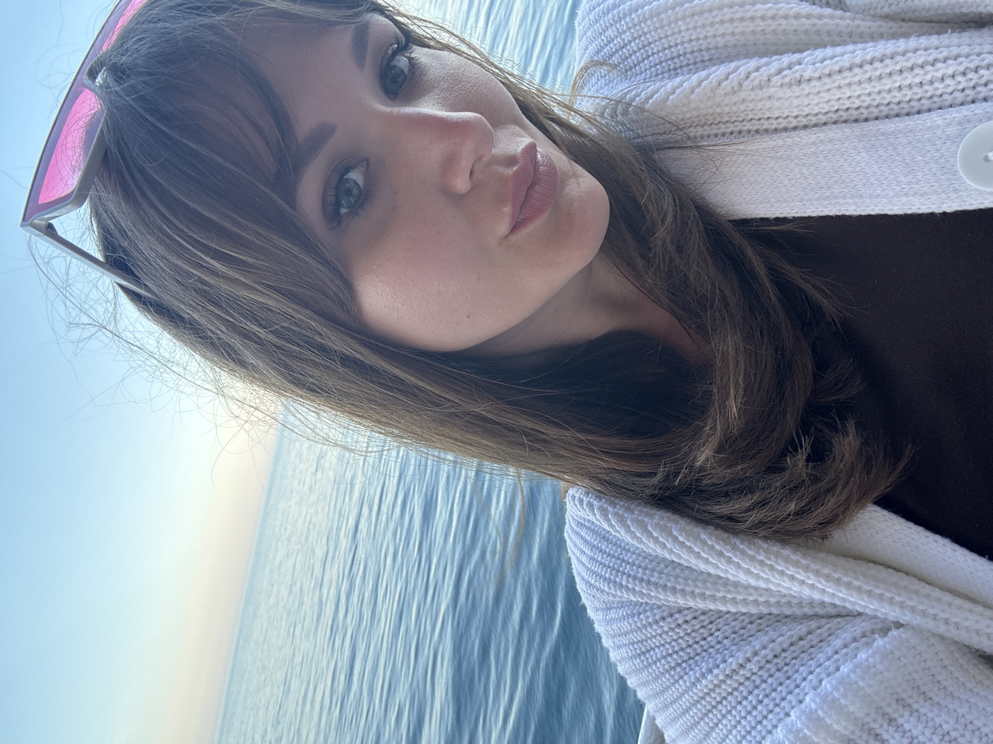 A woman with brown hair and bangs, wearing pink sunglasses, is posing for a selfie. She is puckering her lips. She is wearing a white sweater with a button. Behind her is the sea. The location is likely in or near Гаспринский поселковый совет, Ukraine, given the possible outdoor and sea background.
